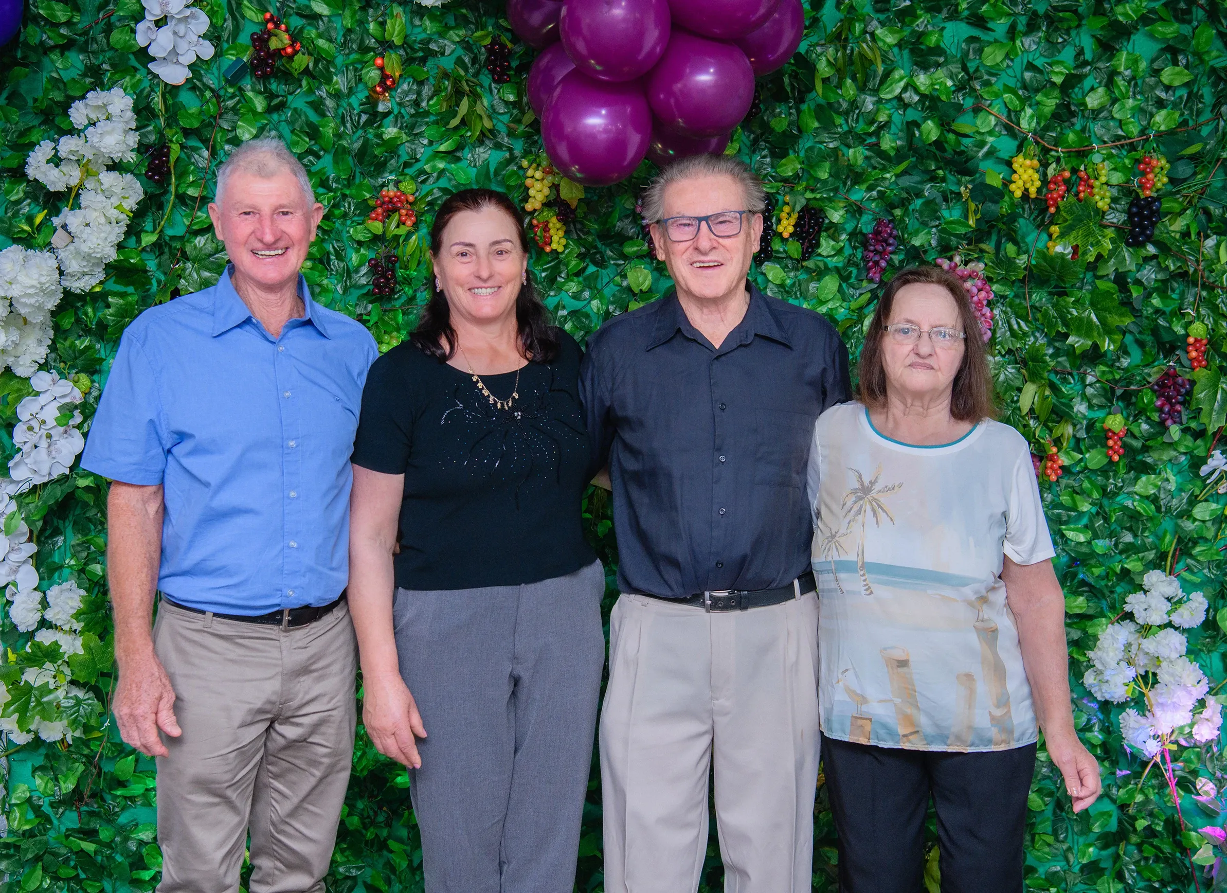 Quatro pessoas adultas, duas mulheres e dois homens, posam em frente a uma parede coberta de hera com cachos de uvas roxas e flores brancas e roxas.