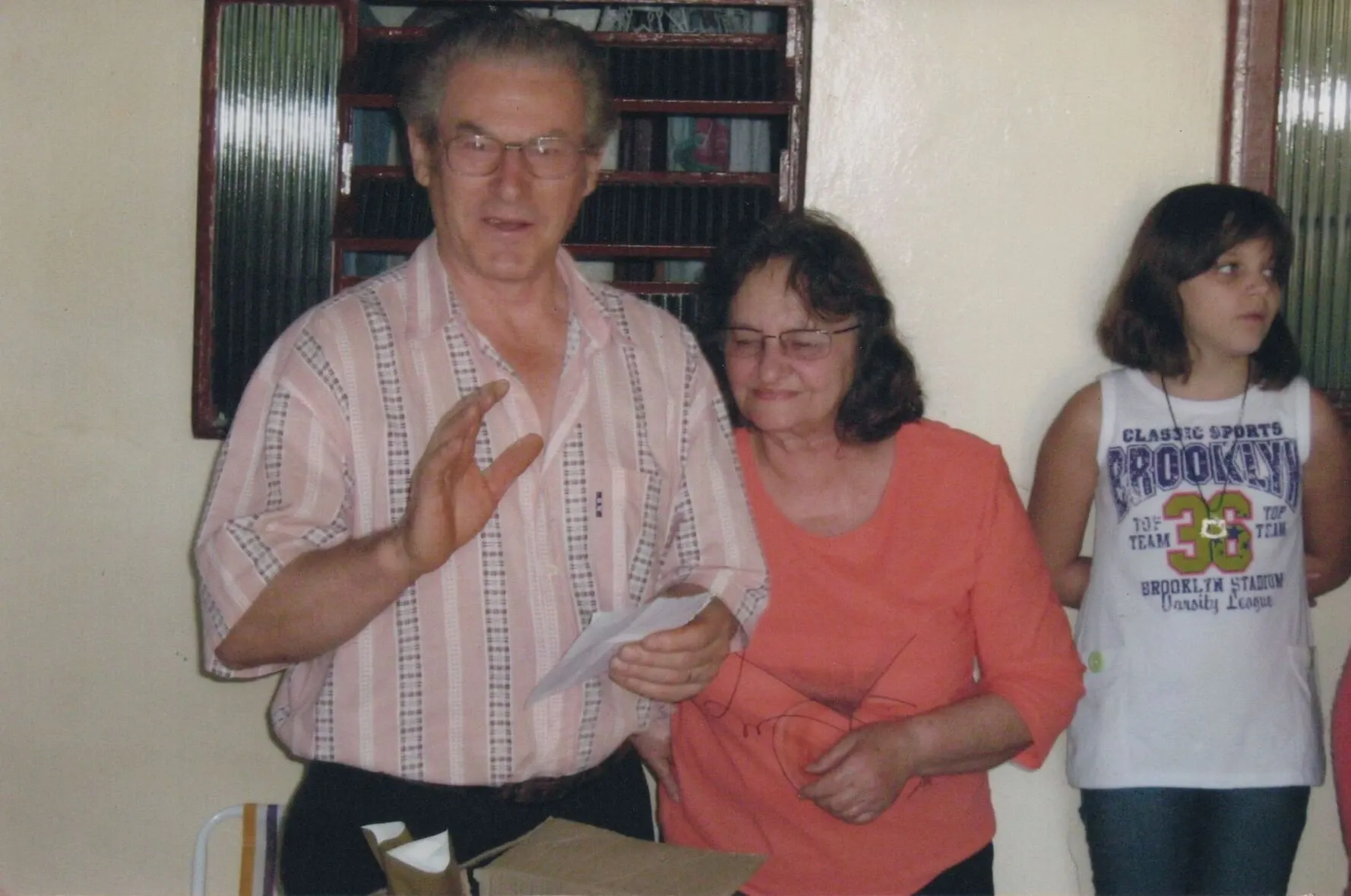 Um homem e uma mulher em primeiro plano, ladeados por uma garota à direita. O homem, com óculos e camisa xadrez, gesticula com a mão direita e segura um papel na esquerda. A mulher, de óculos e blusa laranja, olha para o papel com a mão na cintura do homem. A garota veste uma camiseta branca e olha para a direita.