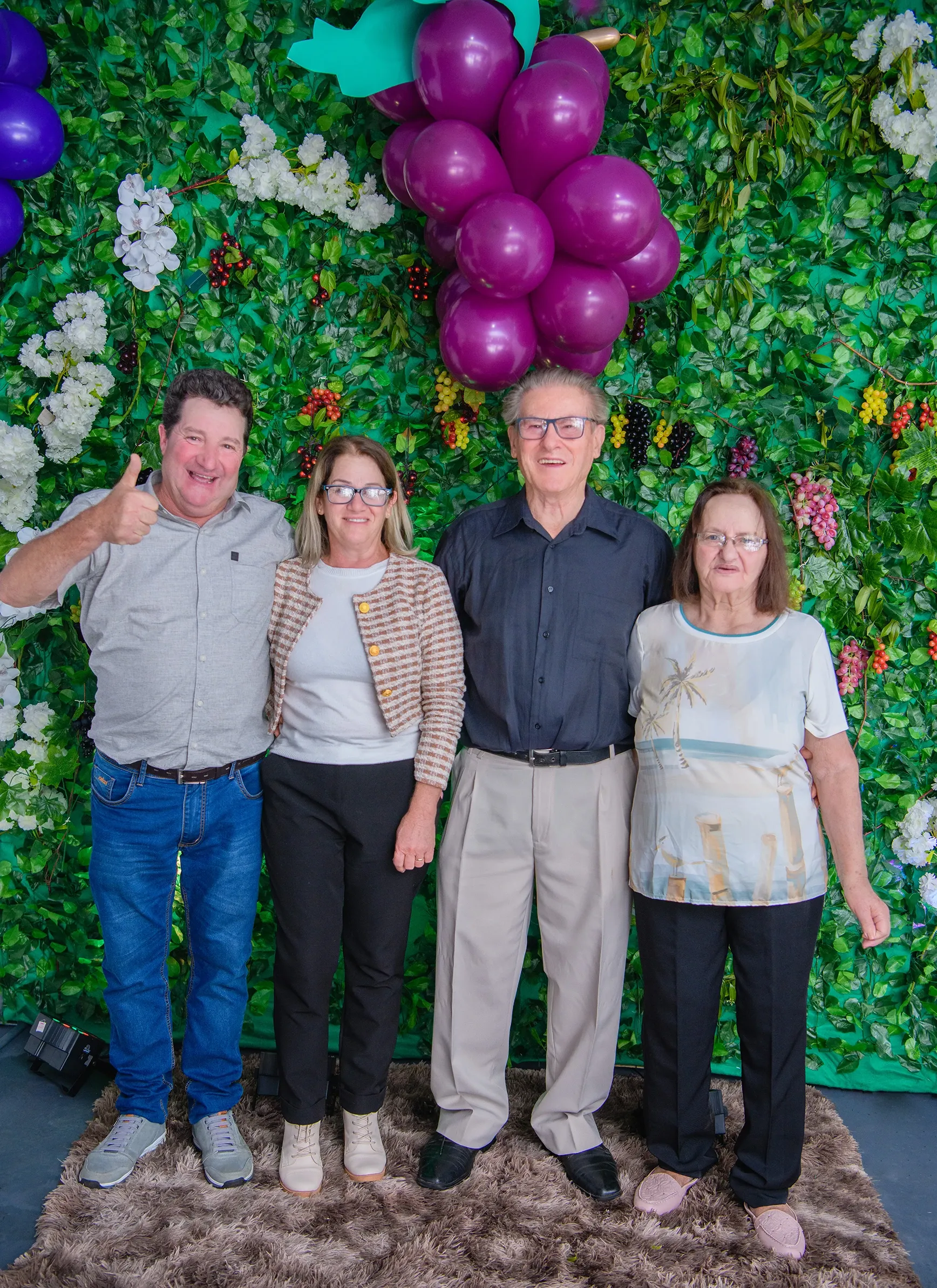 Quatro pessoas (dois homens e duas mulheres) de pé, sorrindo, em frente a uma parede de folhagem verde decorada com flores brancas e roxas e cachos de balões roxos em forma de uva; o homem à esquerda mostra um polegar para cima.