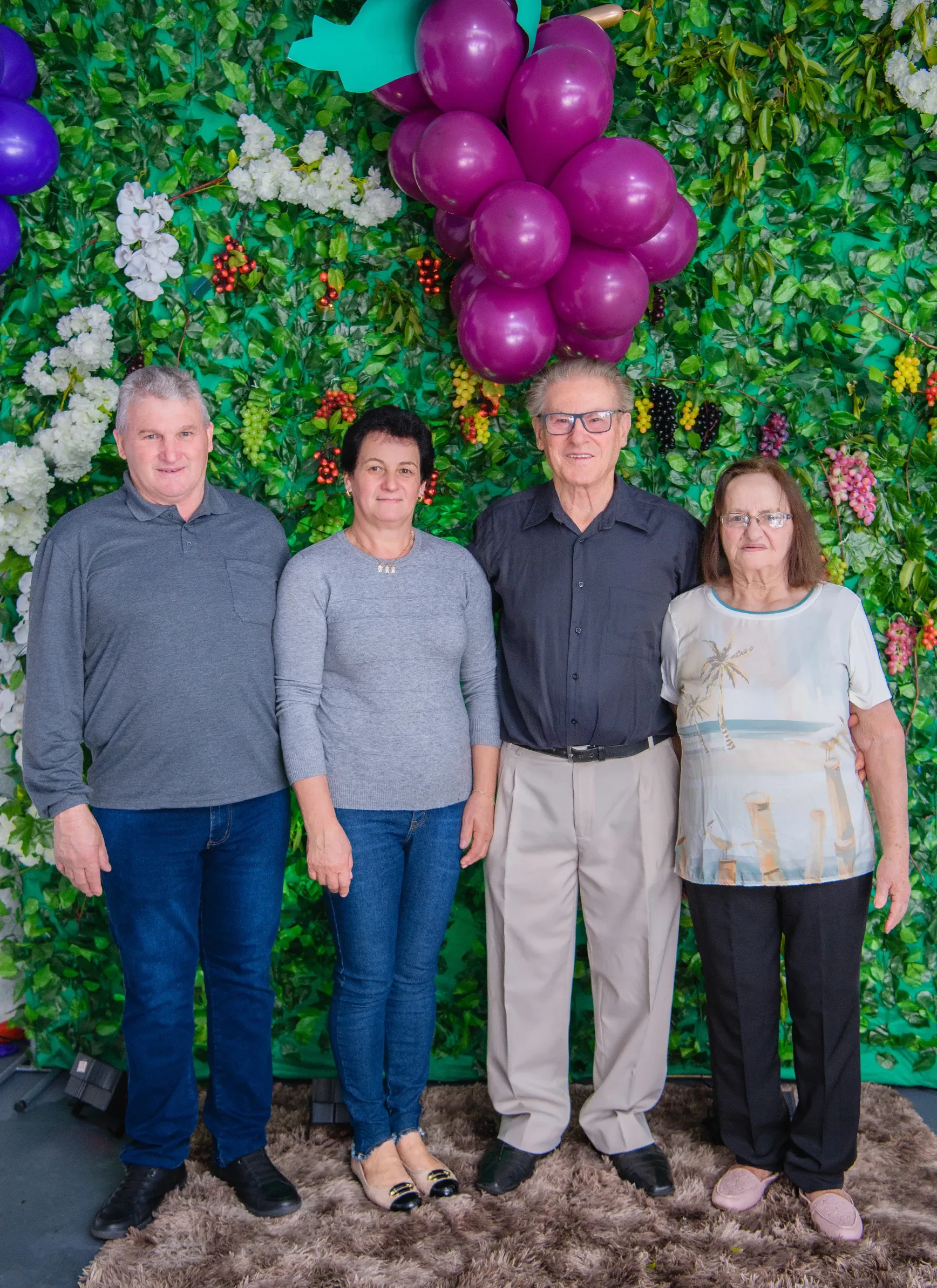Quatro pessoas, dois homens e duas mulheres, estão em pé na frente de uma parede coberta por folhagens verdes e flores brancas, com balões roxos no canto superior direito.