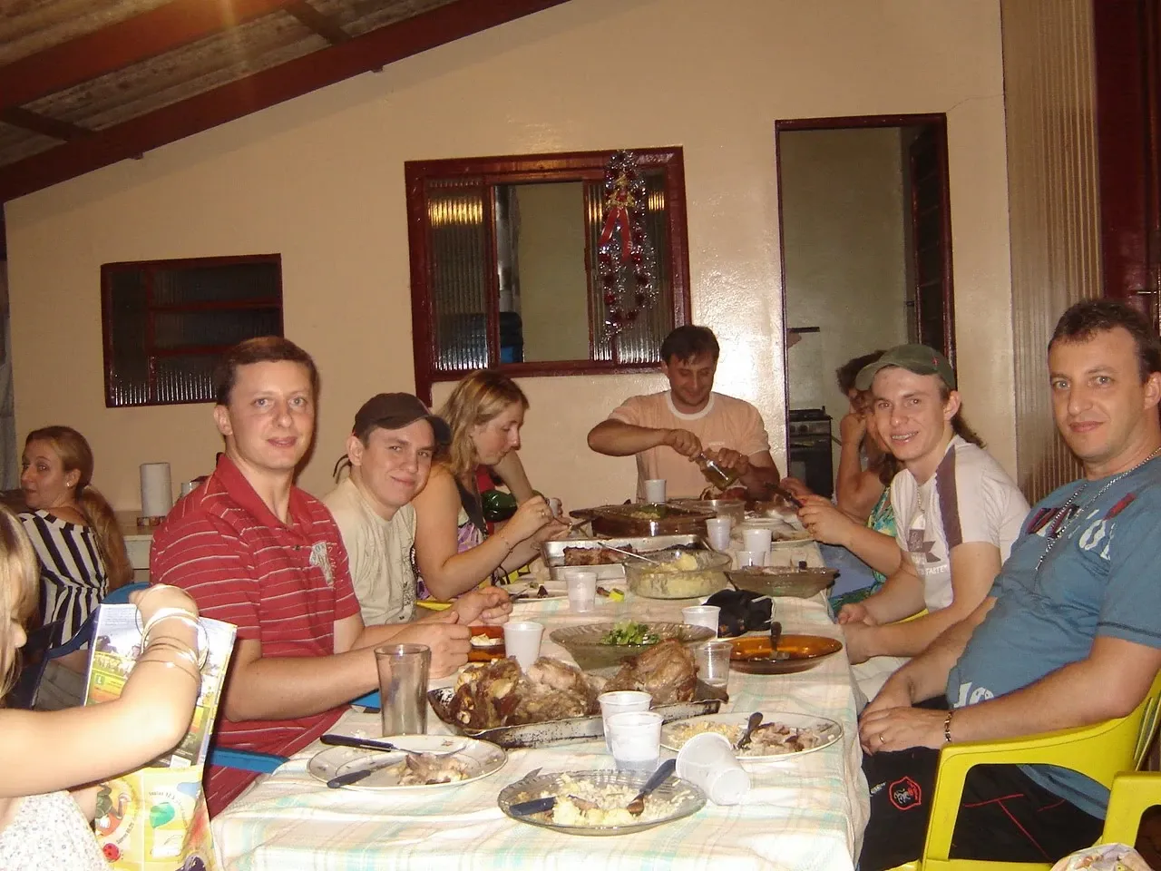 Um grupo de nove pessoas está sentado ao redor de uma mesa de jantar, cheia de comida e pratos, dentro de casa.