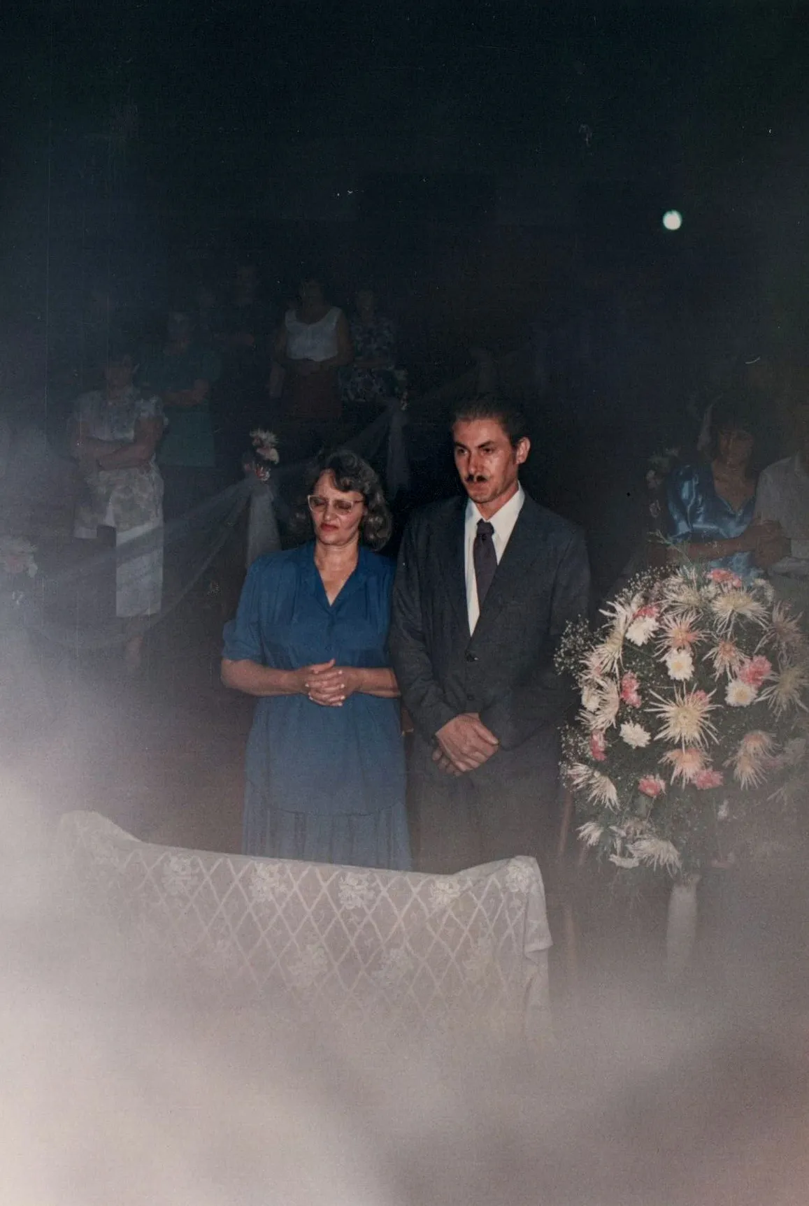 Mulher de blusa azul e homem de terno cinza em pé à beira de um caixão, com arranjos de flores e outras pessoas em segundo plano.