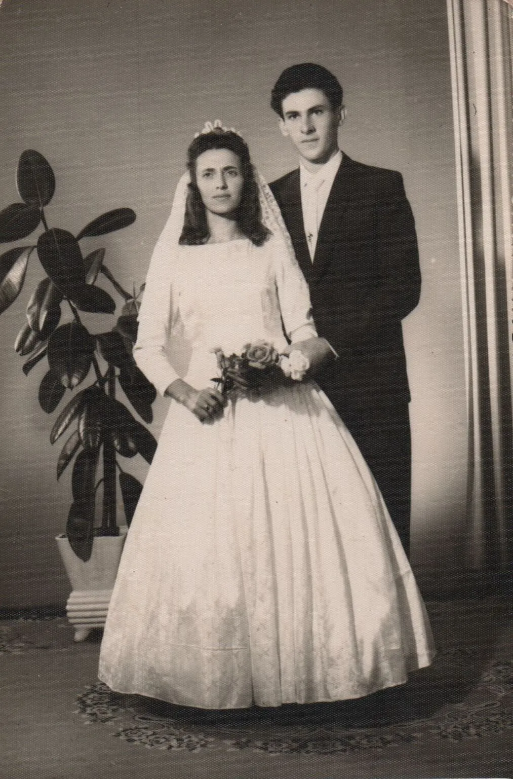 Uma foto de casamento vintage apresenta um casal jovem: a noiva está em primeiro plano, vestindo um vestido de noiva branco e segurando um buquê de rosas, enquanto o noivo está atrás dela, vestindo um terno cinza. (Original)