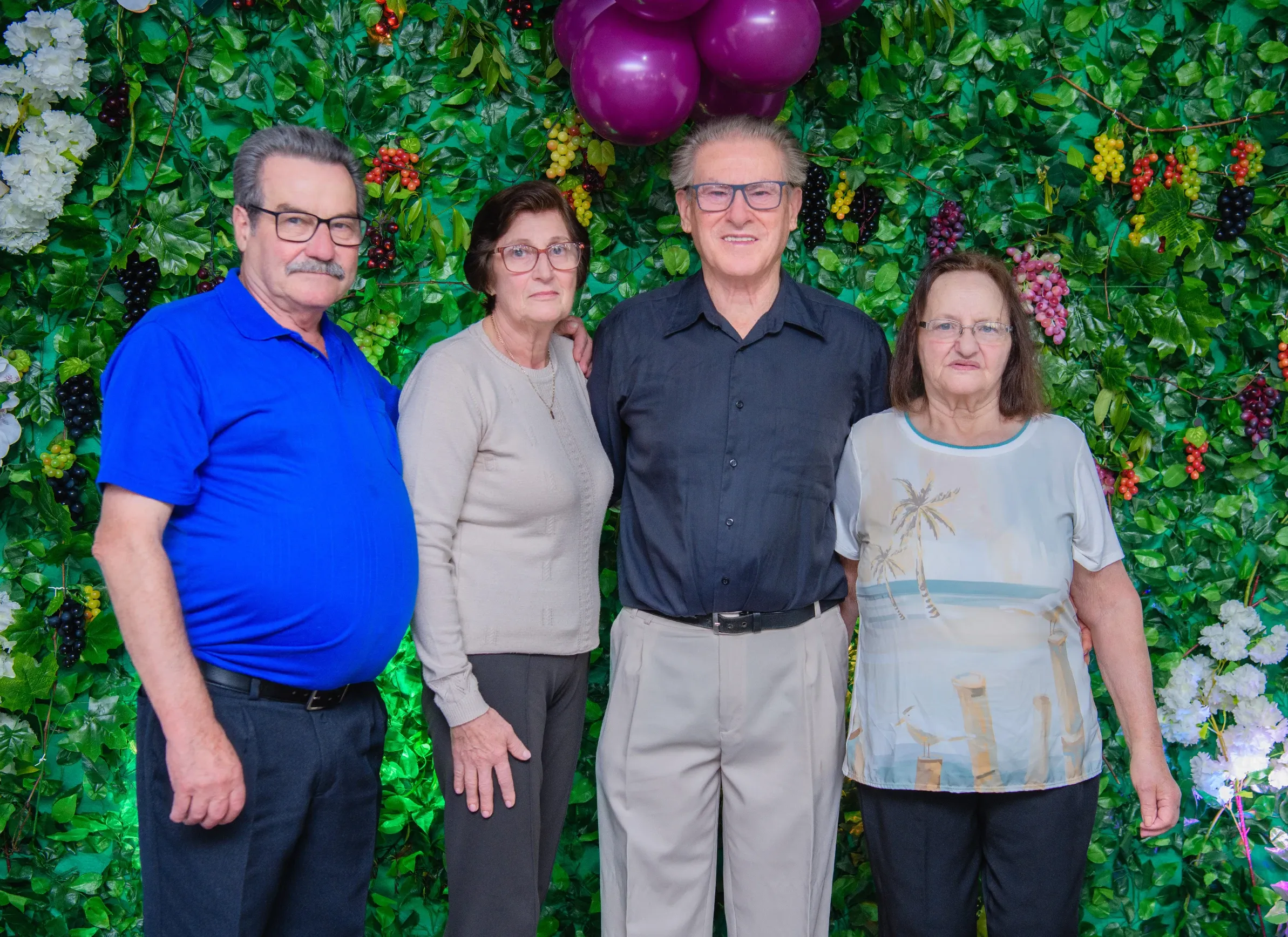 Quatro adultos sorridentes, dois homens e duas mulheres, estão de pé e posando para a câmera. Ao fundo, um arranjo floral em tons de verde e branco é adornado com uvas roxas e escuras. No canto superior direito, um cacho de balões roxos adiciona um toque festivo.