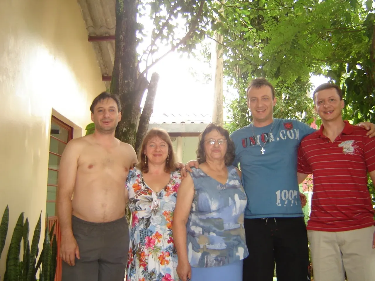 Cinco adultos posam juntos ao ar livre: um homem sem camisa e uma mulher de vestido floral à esquerda, uma mulher de óculos no centro e dois homens com camisetas à direita, em frente a uma parede de cor clara e folhagem verde.