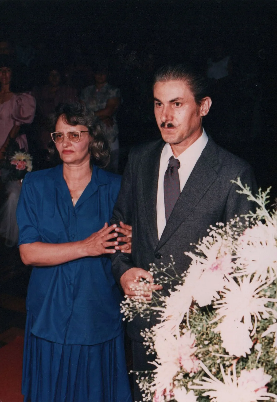 Um homem de terno cinza e bigode estilo dali está ao lado de uma mulher de óculos e blusa azul, segurando um grande buquê de flores brancas.