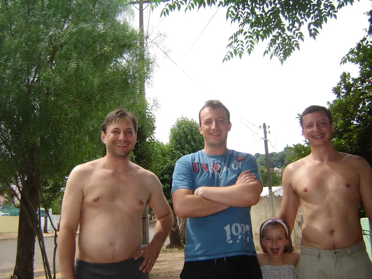 Três homens e uma menina, em frente a uma casa de campo, um deles sorri e abraça a menina, enquanto os outros dois sorriem e posam.