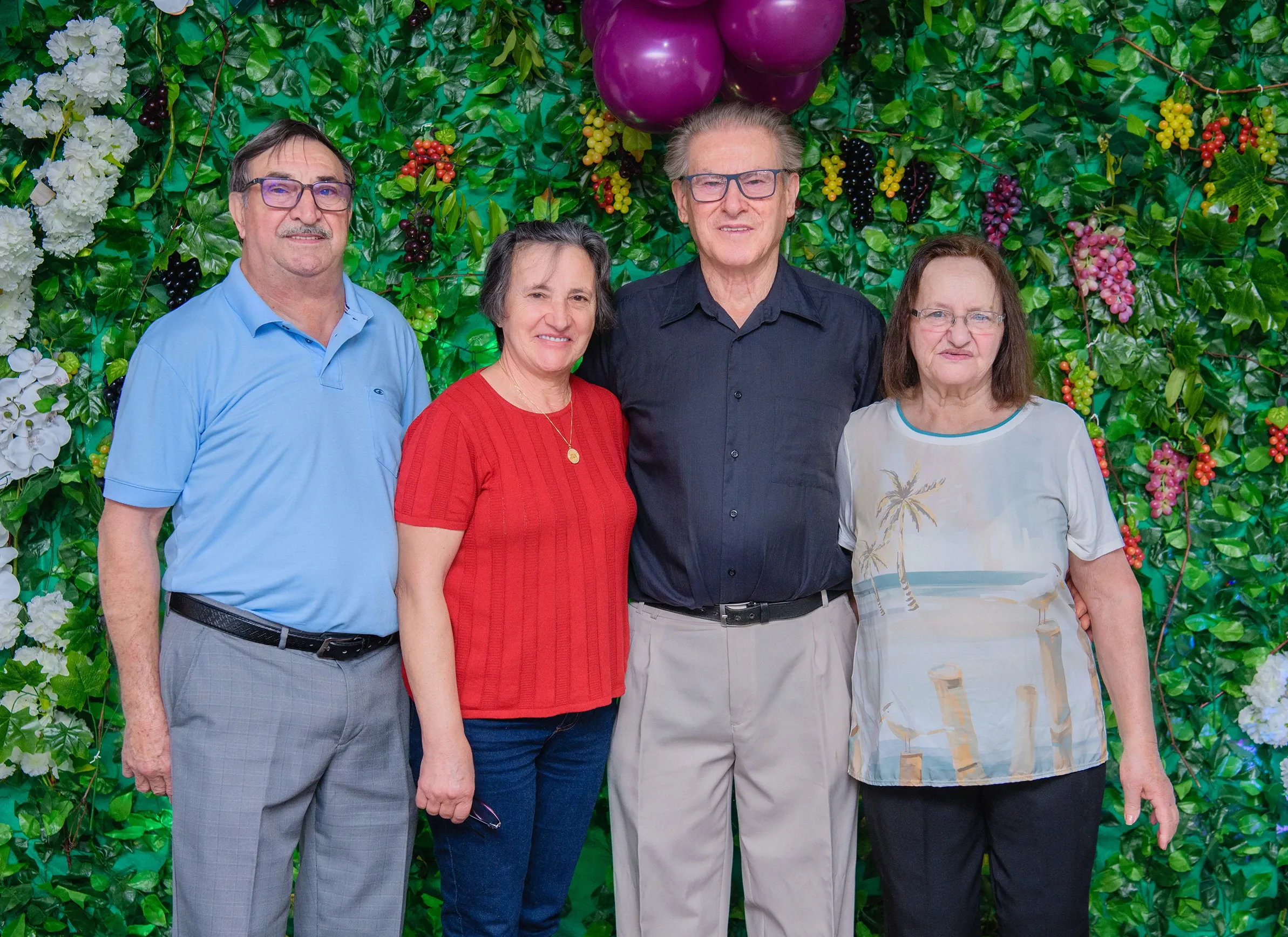 Quatro adultos, dois homens e duas mulheres, estão lado a lado, sorrindo em frente a uma parede de folhagem verde com cachos de uvas roxas e vermelhas e flores brancas.