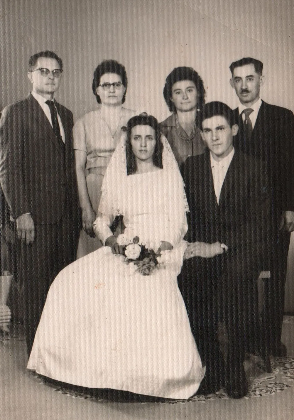 Retrato de família de casamento da década de 1950 com a noiva sentada na frente em um vestido branco e véu, segurando um buquê de rosas brancas, ladeada pelo noivo sentado e outras quatro pessoas em pé atrás. (Original)