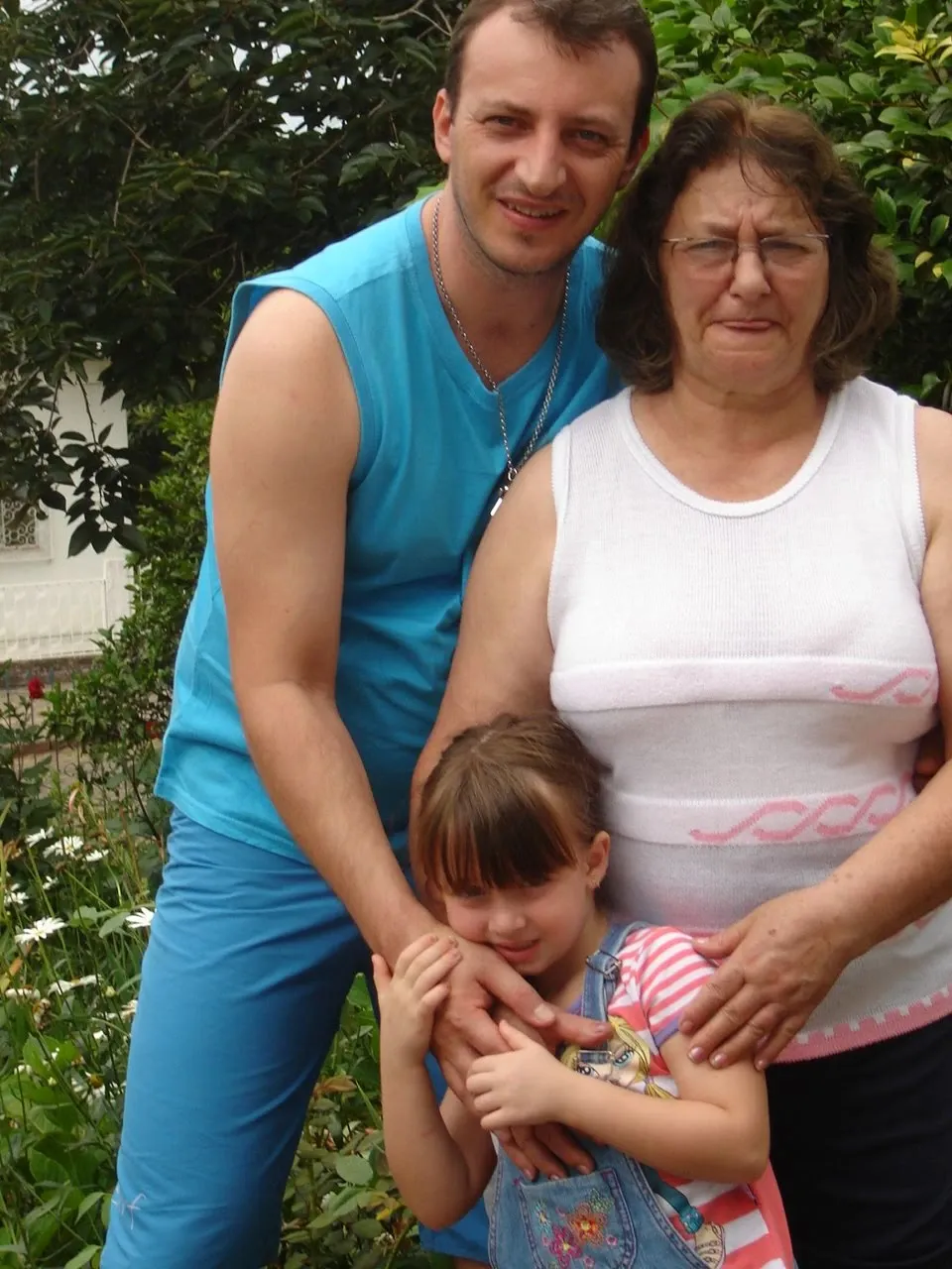 Homem, mulher e menina estão no centro de uma imagem. O homem é alto e está usando um colete azul, uma corrente de prata e calças azuis. Ele tem cabelo castanho escuro e uma barba por fazer. A mulher tem cabelo castanho escuro, usa óculos e uma blusa branca. A menina usa um macacão e uma camiseta listrada. Ela tem cabelo castanho claro e está abraçando as pernas do homem. Os três estão sorrindo, e o plano de fundo é de folhas verdes.