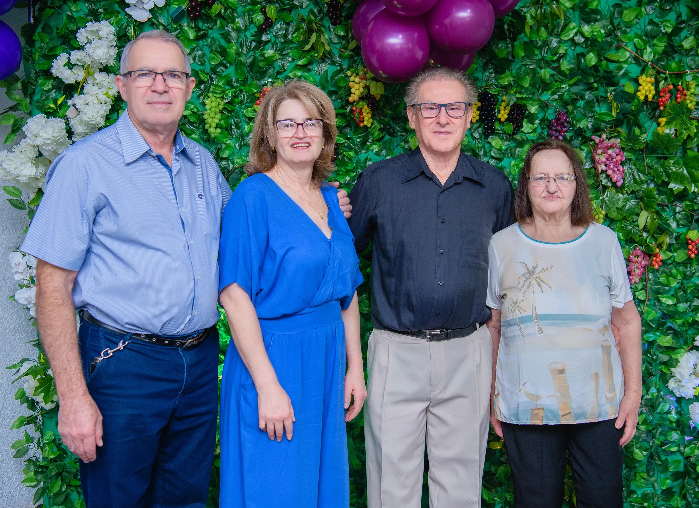 Quatro pessoas sorridentes, dois homens e duas mulheres, em pé na frente de uma parede coberta por folhagem verde e cachos de uva.