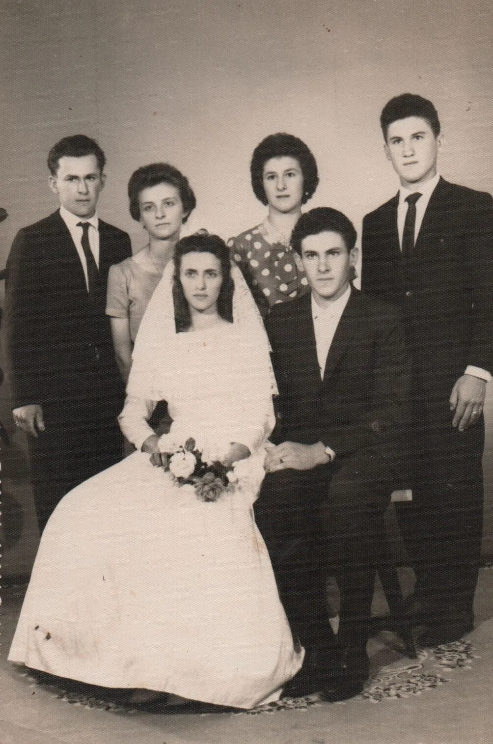 Retrato de estúdio vintage de um casamento com seis pessoas: a noiva e o noivo sentados na frente, a noiva segurando um buquê de rosas, e quatro outros convidados em pé atrás deles. (Original)