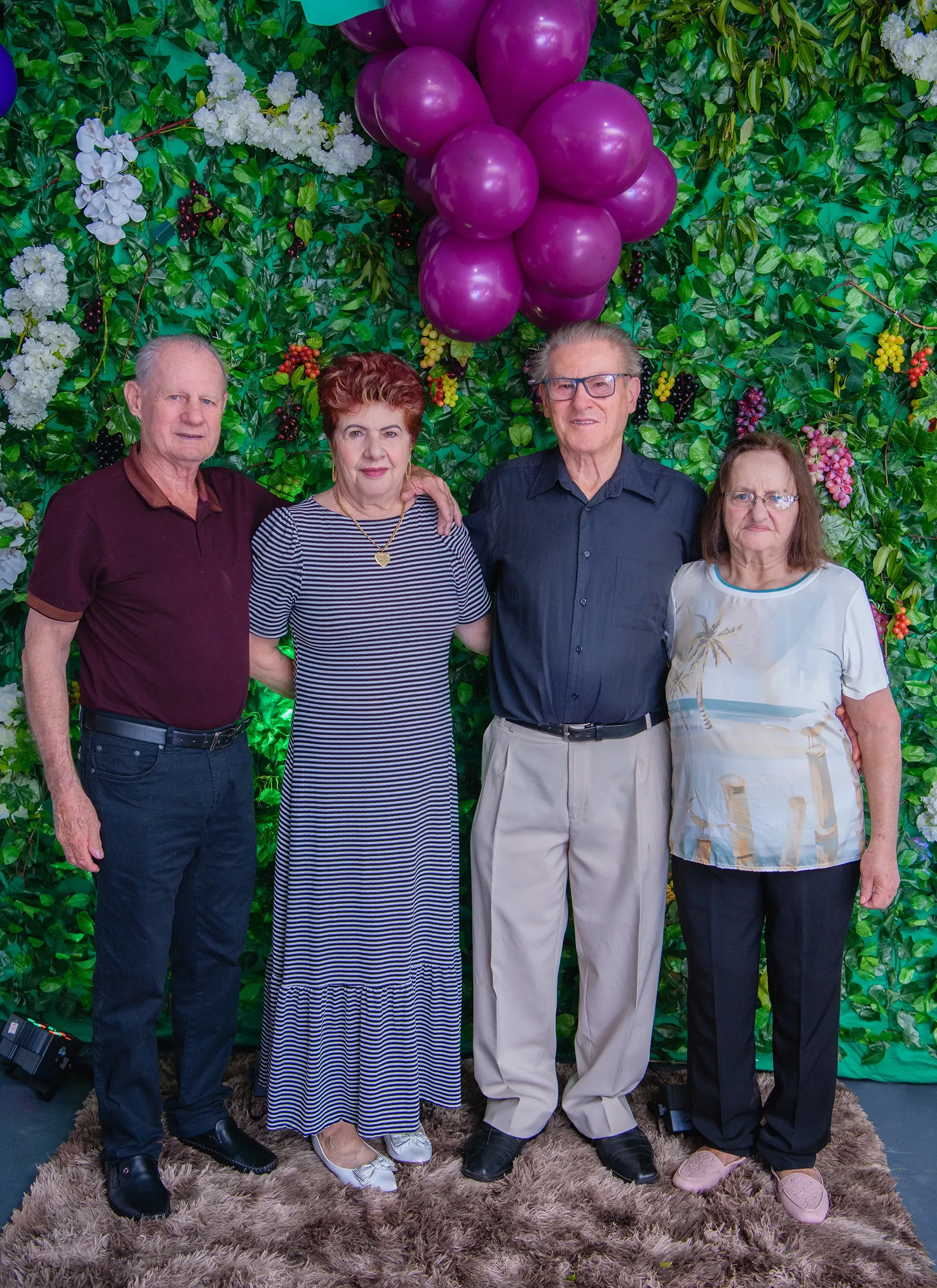Quatro adultos mais velhos, dois homens e duas mulheres, estão de pé e sorrindo, abraçando-se, na frente de uma parede de folhagem verde adornada com flores brancas e roxas, frutas escuras e cachos de balões roxos no canto superior direito.