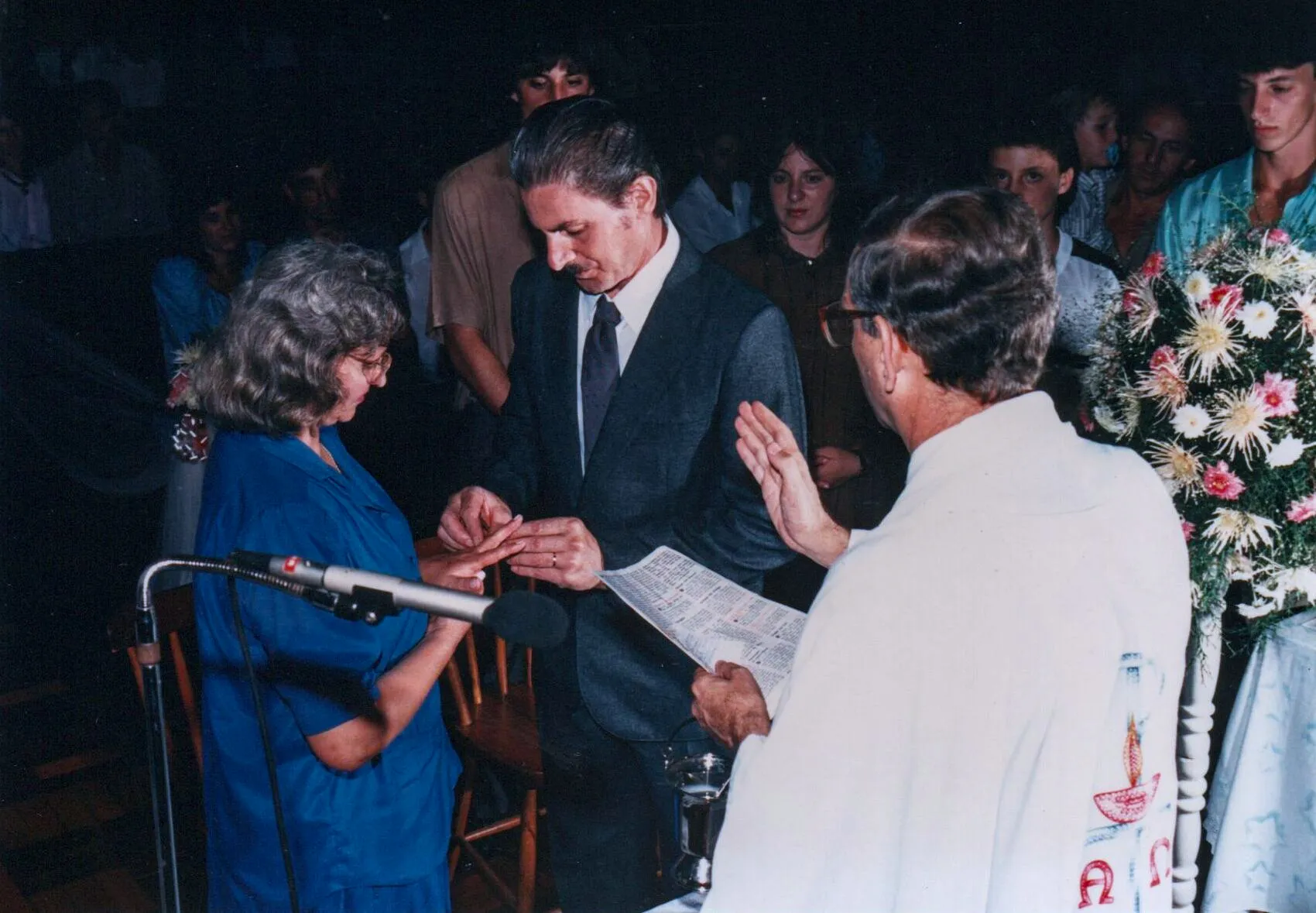 Um homem de terno colocando um anel no dedo de uma mulher de blusa azul durante uma cerimônia de casamento, com um sacerdote de túnica branca à direita e convidados ao fundo.