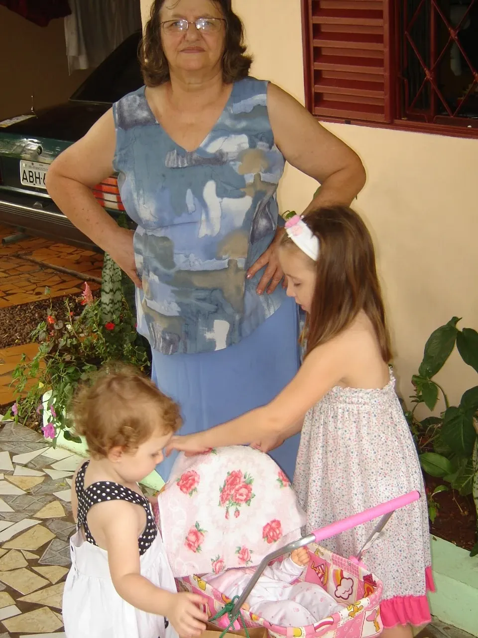 Uma mulher e duas meninas em um quintal. A mulher, em pé, olha para frente enquanto as duas meninas brincam com um carrinho de bebê de brinquedo. A menina mais velha, de vestido florido, empurra o carrinho enquanto a mais nova, de vestido de bolinhas, olha para dentro dele.