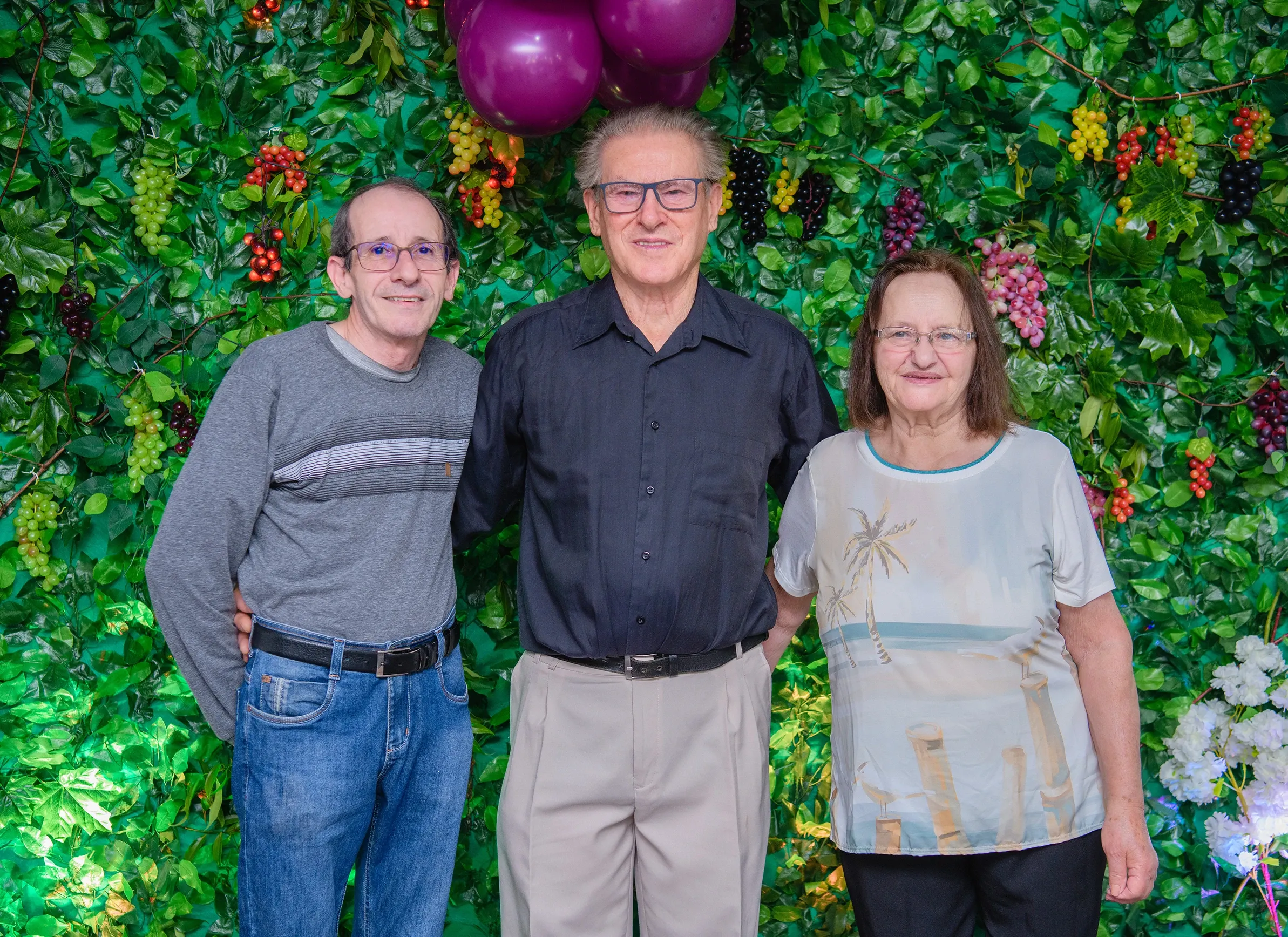 Três pessoas sorridentes — dois homens e uma mulher — posam em frente a uma parede de folhagem verde adornada com uvas artificiais e balões roxos.