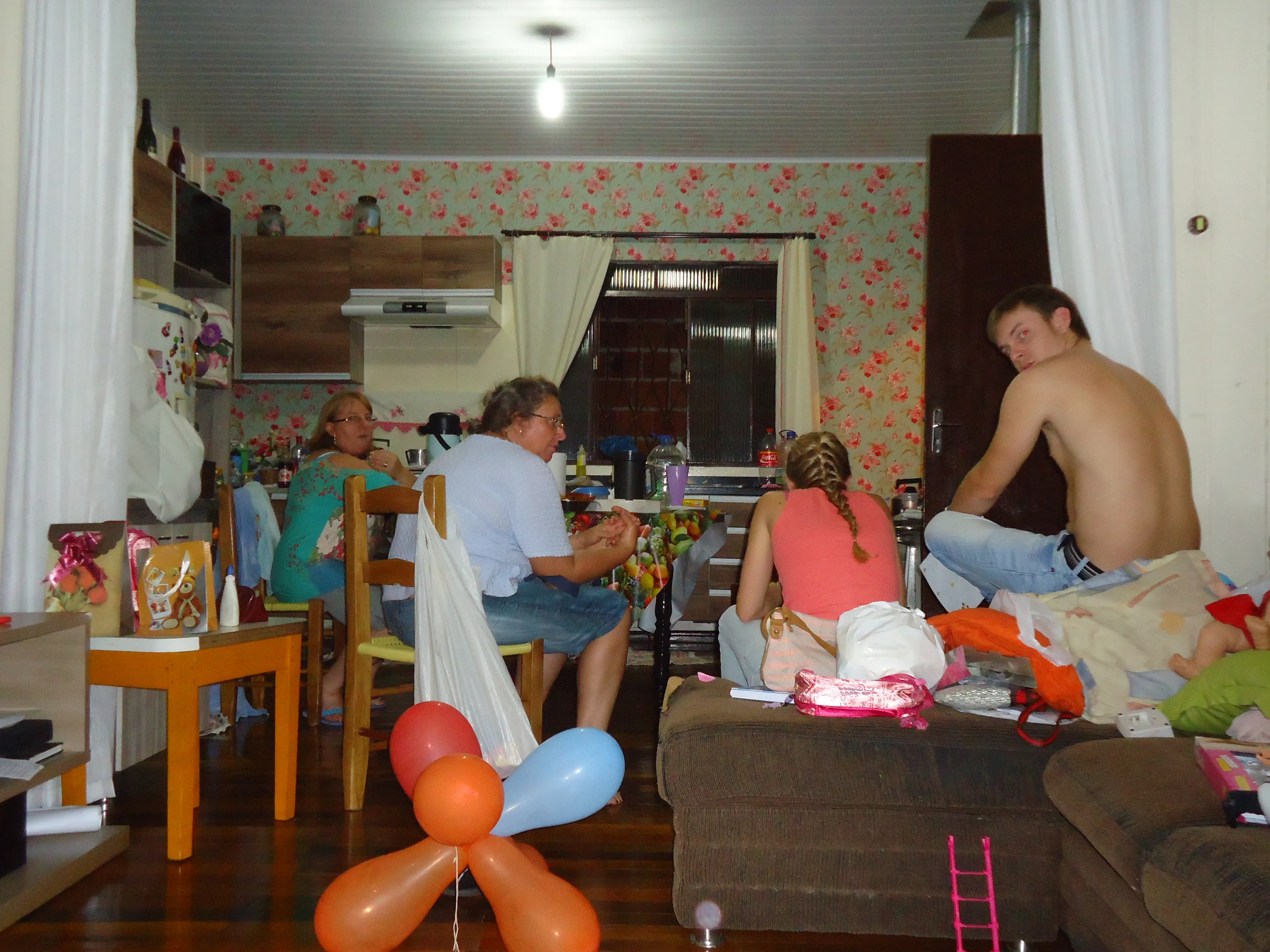Um grupo de cinco pessoas está relaxando em uma sala de estar com uma decoração floral, com três sentadas à mesa e duas sentadas em um sofá, enquanto balões laranja e azuis flutuam no chão.