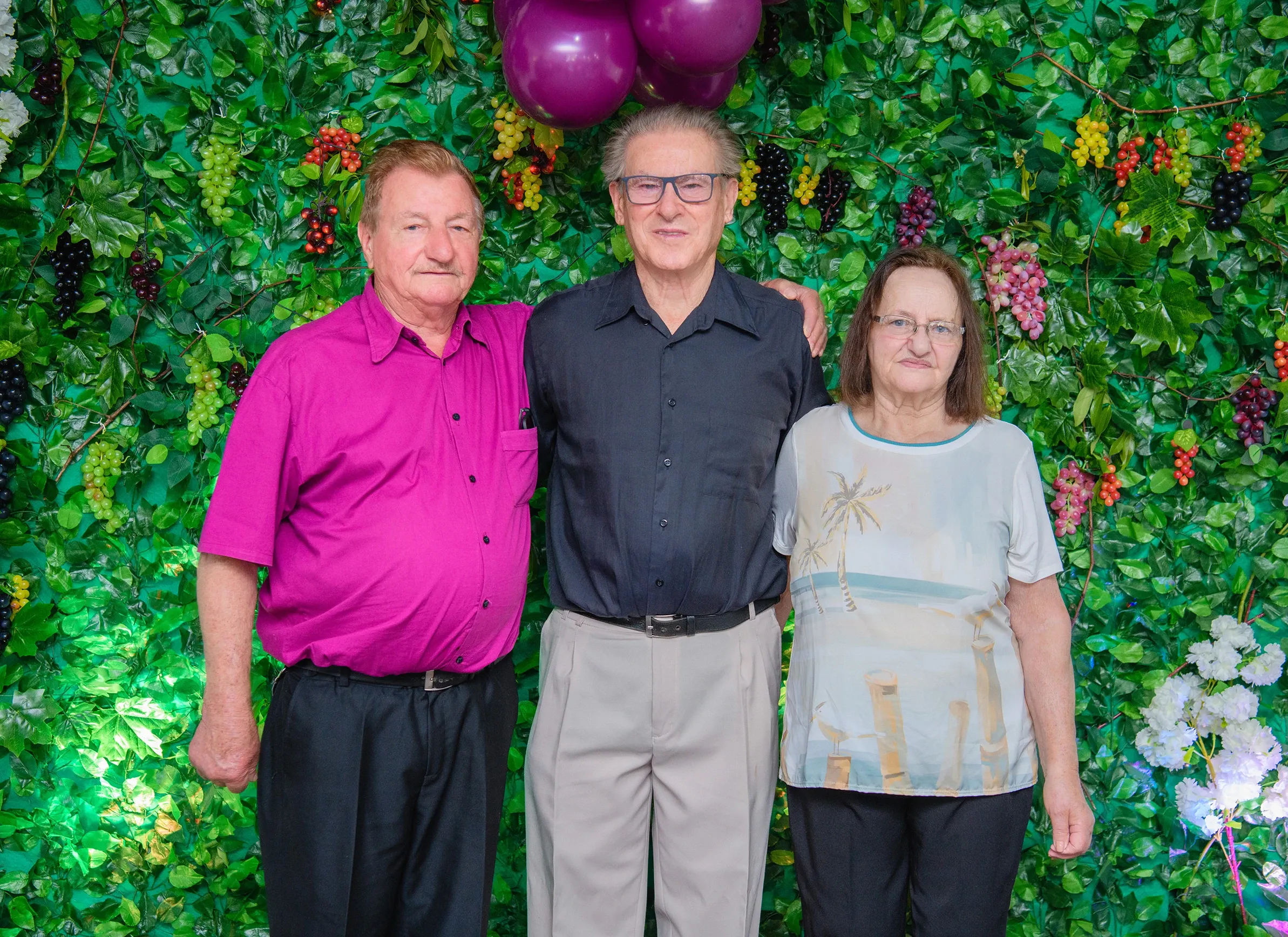 Três pessoas idosas, dois homens e uma mulher, sorriem e se abraçam em frente a um jardim vertical exuberante com uvas, balões roxos e flores brancas.
