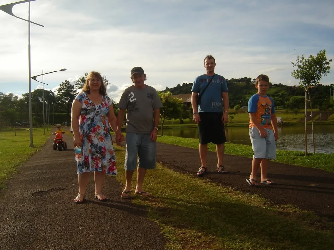 Quatro pessoas — uma mulher, dois homens e um menino — posam em uma estrada de asfalto ao lado de um lago, com grama verde e árvores em um dia claro.
