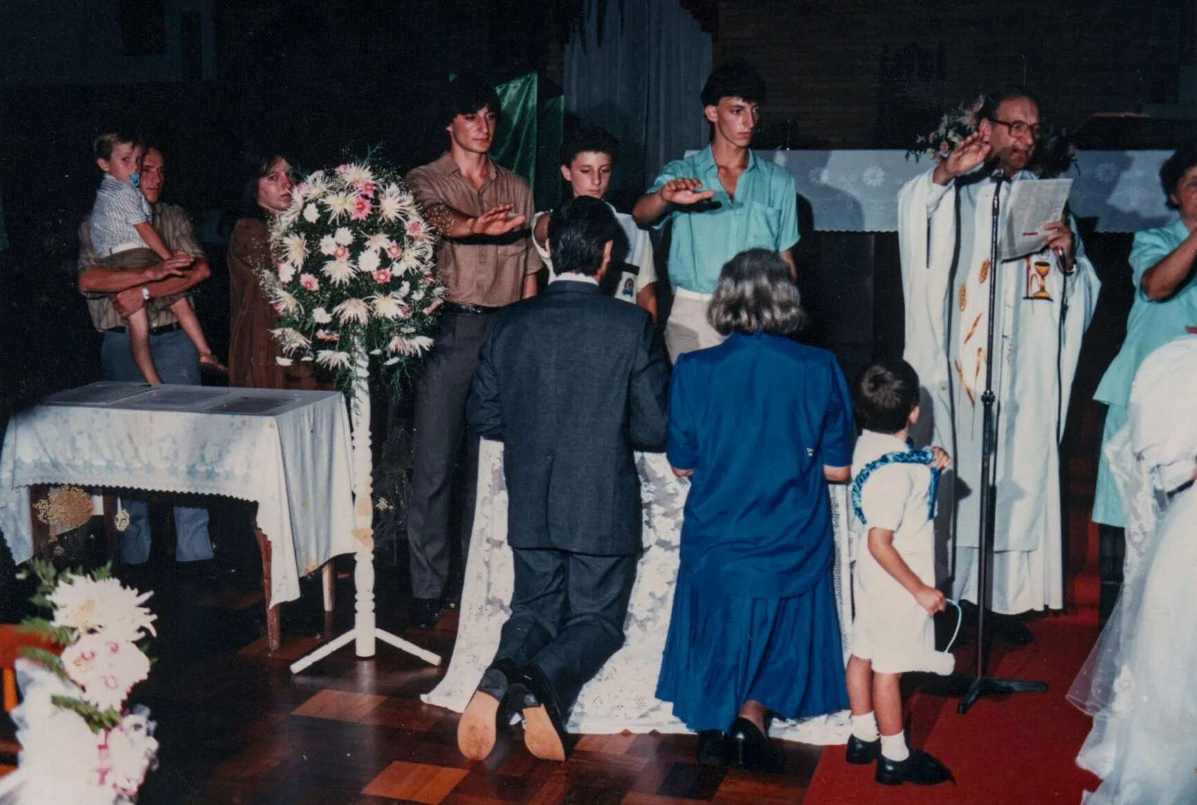 Homem e mulher ajoelhados na frente de um altar em uma cerimônia de casamento, com um padre e convidados assistindo.