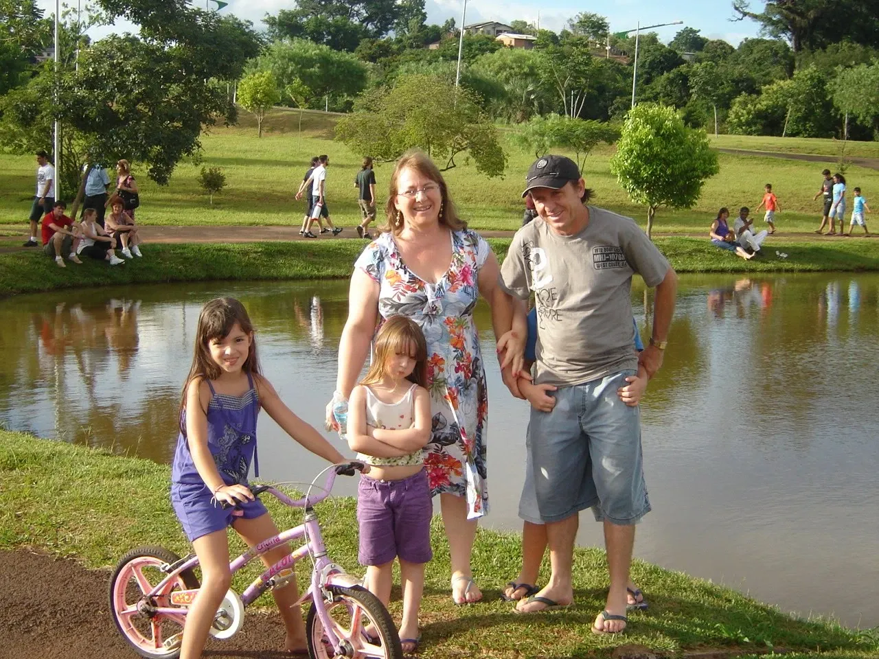 Uma família de quatro pessoas, incluindo duas crianças e dois adultos, posa em frente a um lago em um parque ensolarado.