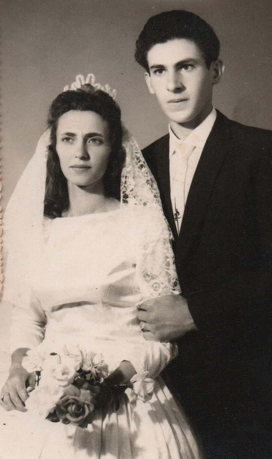 Uma jovem noiva em um vestido de cetim branco com buquê de flores e um noivo de terno preto posam para uma foto de casamento vintage. (Original)