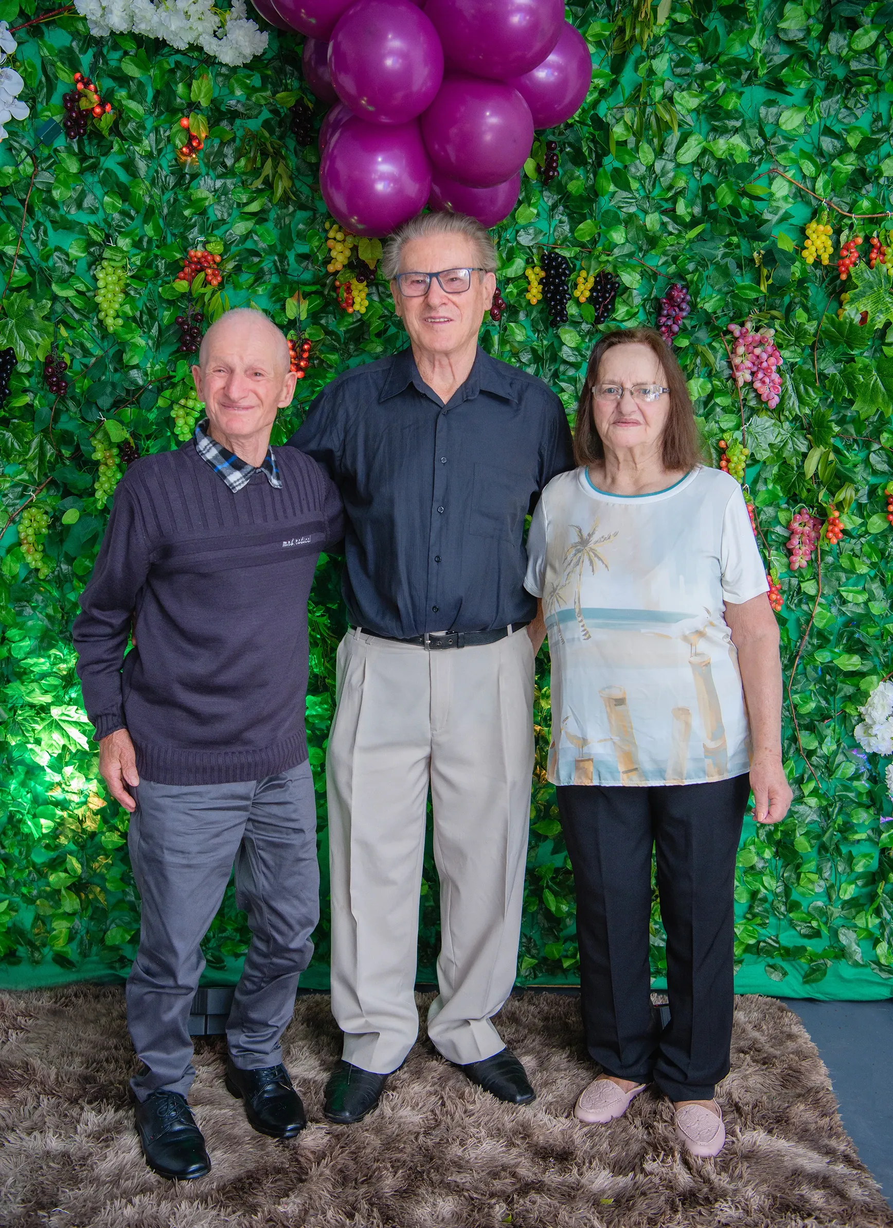 Três idosos, dois homens e uma mulher, em pé na frente de uma parede coberta de folhagem verde e cachos de frutas e balões roxos. O homem à esquerda usa uma blusa de lã azul-marinho, calças cinzas e sapatos pretos. O homem no centro usa uma camisa preta, calças cáqui e sapatos pretos. A mulher à direita usa uma camiseta branca com estampa de palmeira, calças pretas e sapatos rosa. Todos sorriem para a câmera.