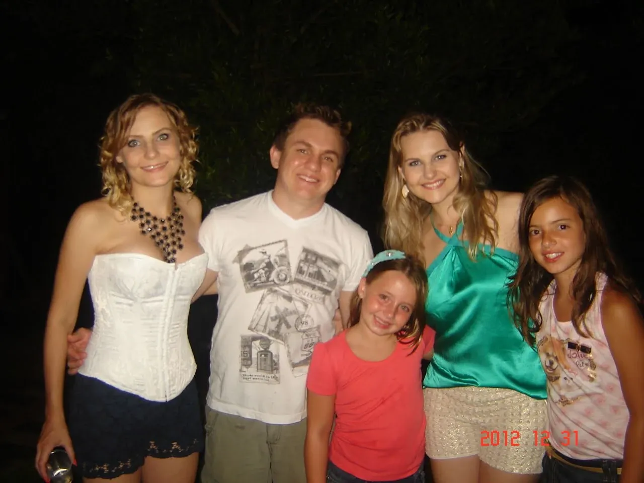 Cinco pessoas, incluindo três mulheres adultas e duas meninas, posam ao ar livre à noite com um fundo escuro, todos sorrindo e olhando para a câmera.