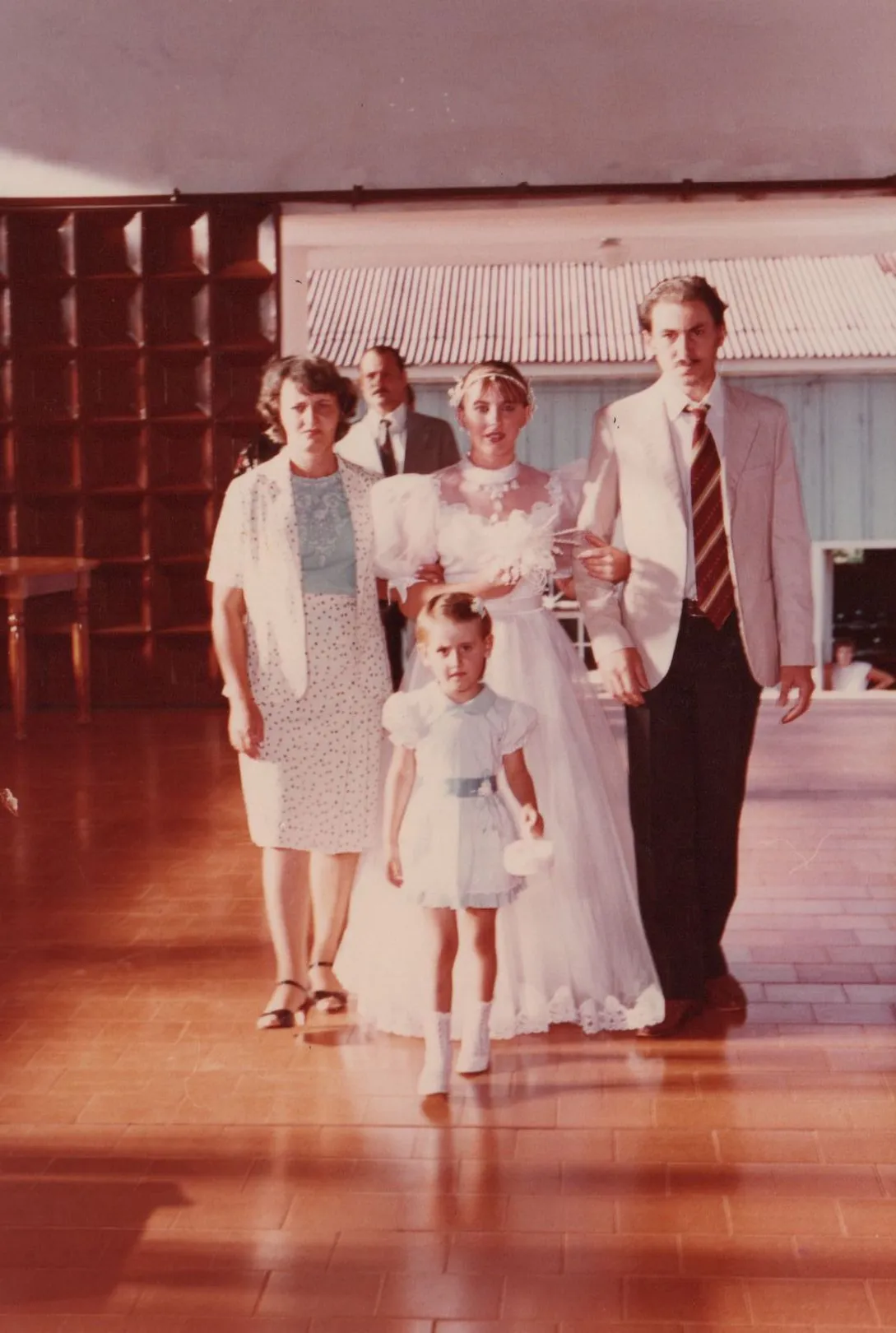 Uma noiva de branco com seu pai e mãe, e uma florista caminhando em um corredor. (Original)