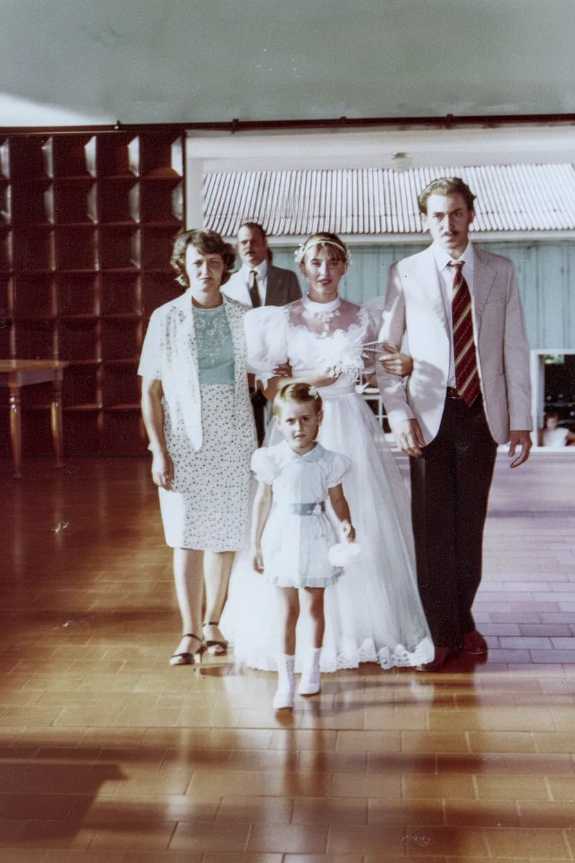 Uma noiva de branco com seu pai e mãe, e uma florista caminhando em um corredor.
