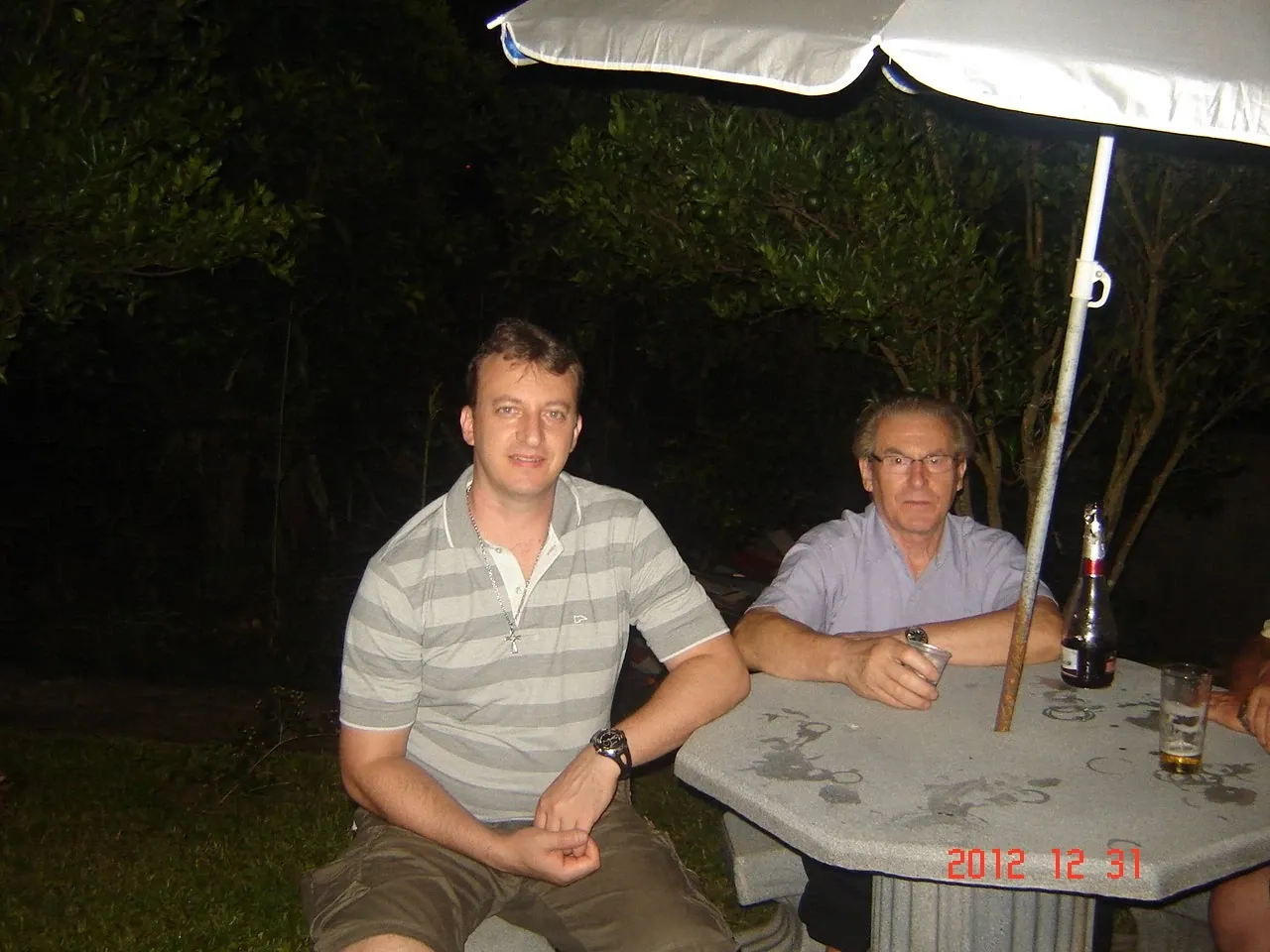 Dois homens, um jovem e um mais velho, sentados à noite sob um guarda-chuva em uma mesa de pátio externa com bebidas.