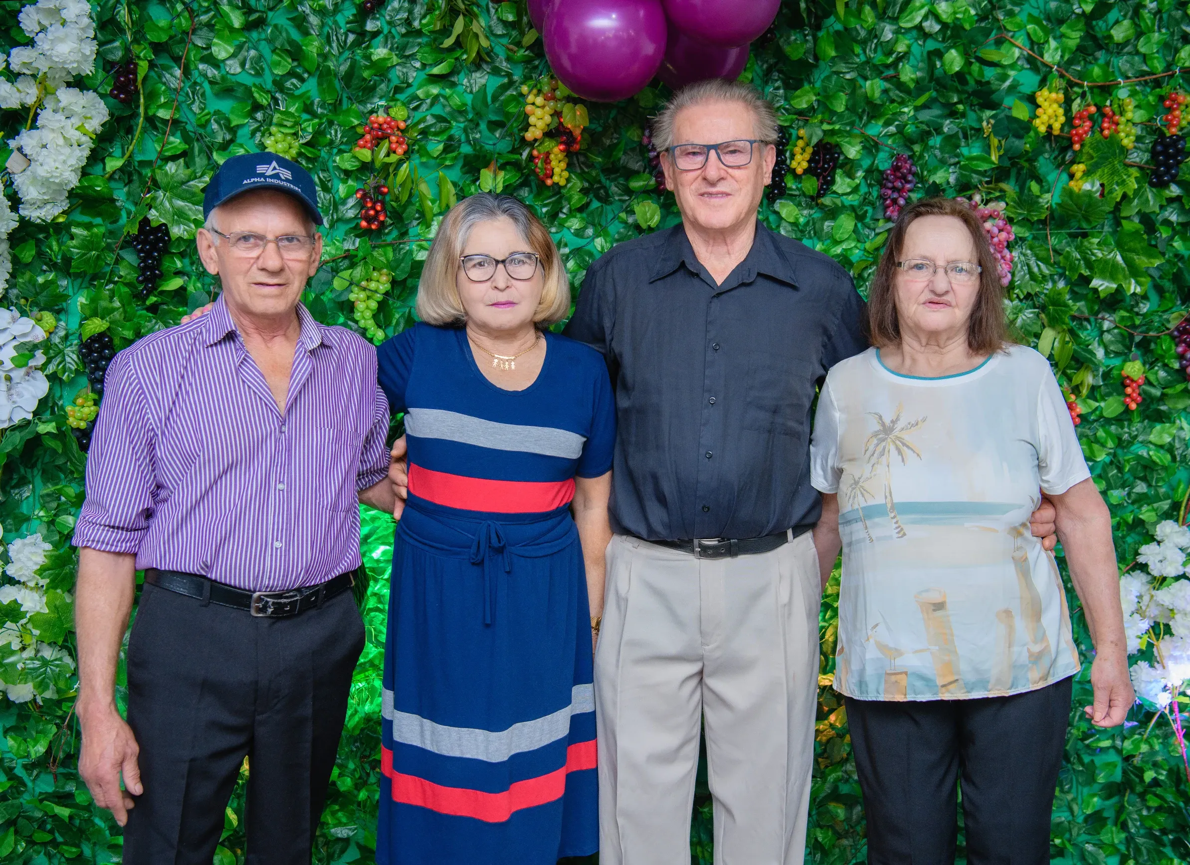 Quatro adultos em pé na frente de uma parede verde coberta de folhas e frutas roxas e vermelhas.