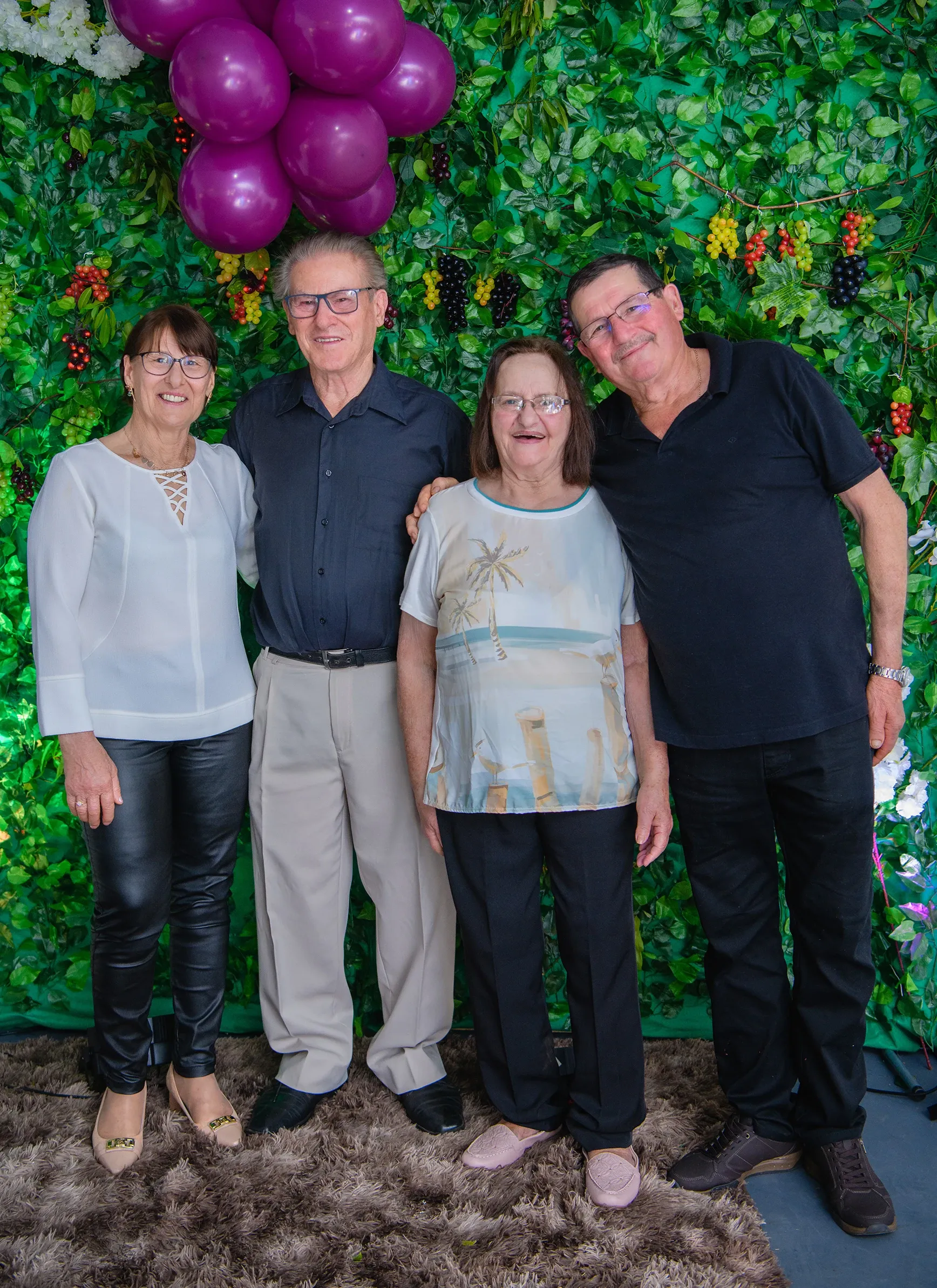 Quatro pessoas posam na frente de uma parede coberta de plantas e frutas, com balões roxos no canto superior esquerdo.