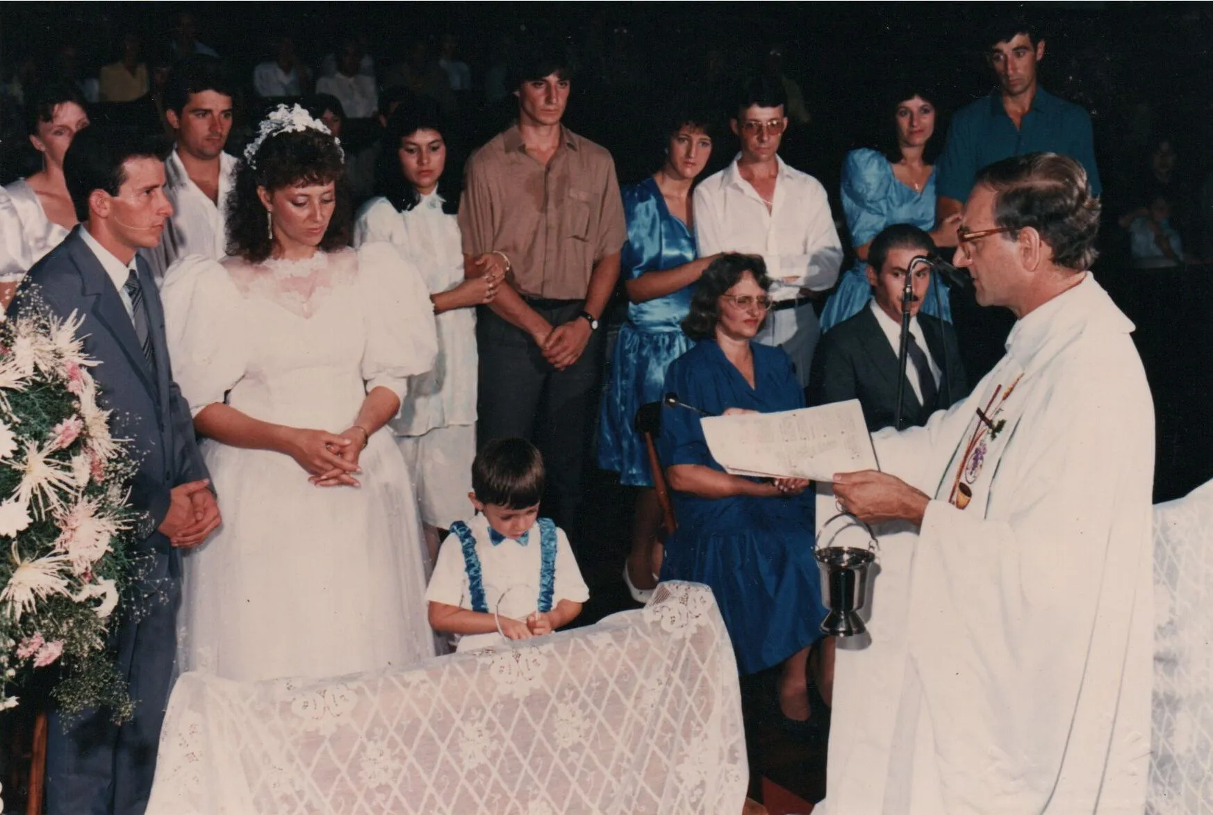 Uma cerimônia de batismo dentro de uma igreja, com uma mulher de vestido de noiva ao lado de um homem de terno, com um padre batizando uma criança pequena, tudo em frente a uma congregação. (Original)