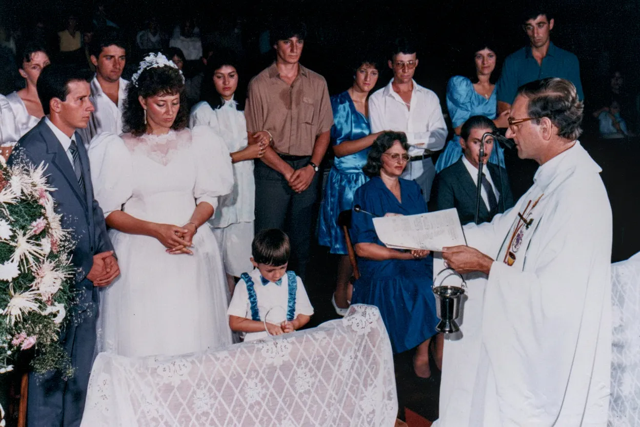 Uma cerimônia de batismo dentro de uma igreja, com uma mulher de vestido de noiva ao lado de um homem de terno, com um padre batizando uma criança pequena, tudo em frente a uma congregação.