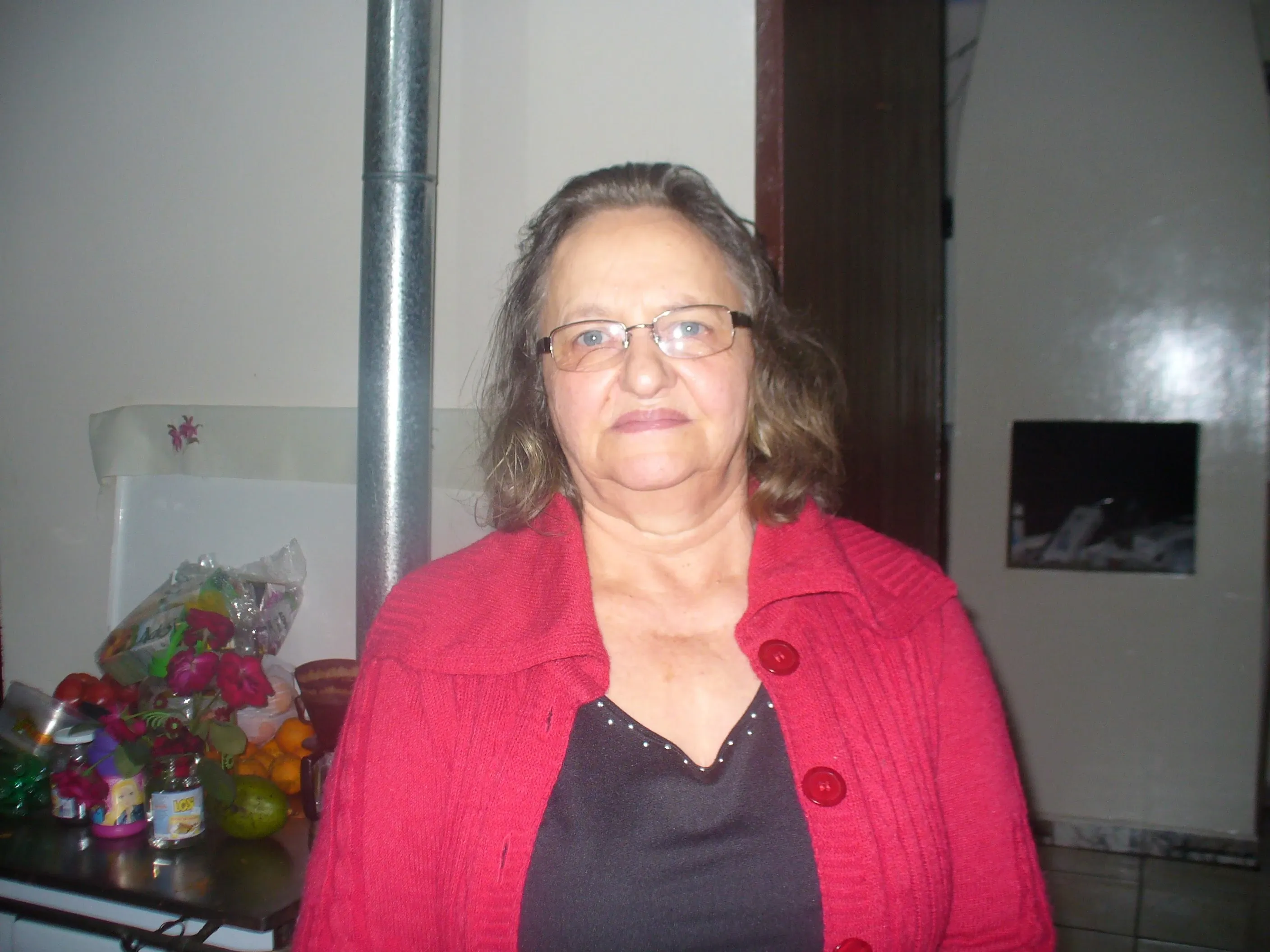 Mulher de meia-idade com óculos e cabelo castanho, vestindo um cardigã vermelho e uma blusa preta, sorri gentilmente para a câmera em uma cozinha iluminada.