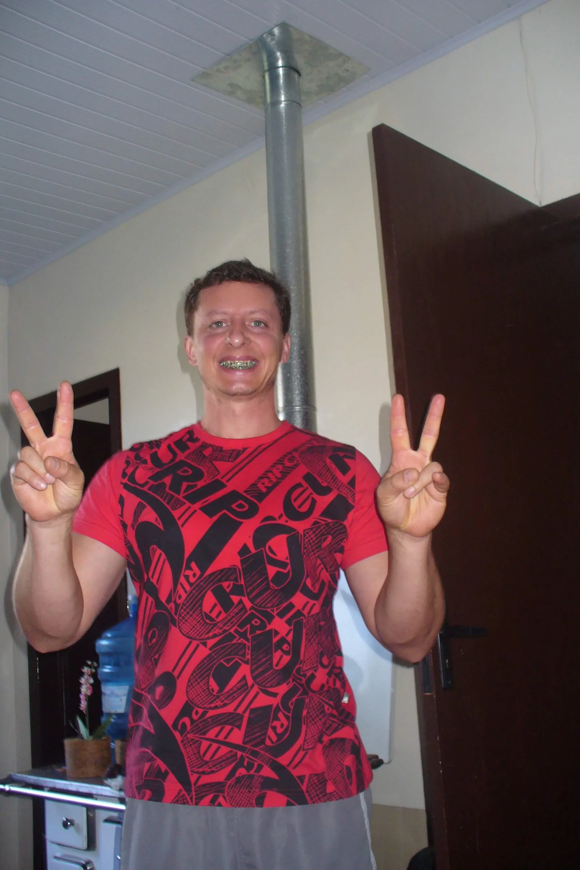 Um homem branco com aparelho nos dentes, usando uma camiseta vermelha estampada e shorts cinzas, sorri e faz um sinal de "paz" com as duas mãos, em pé em um quarto com uma porta marrom e um cano de metal no teto.
