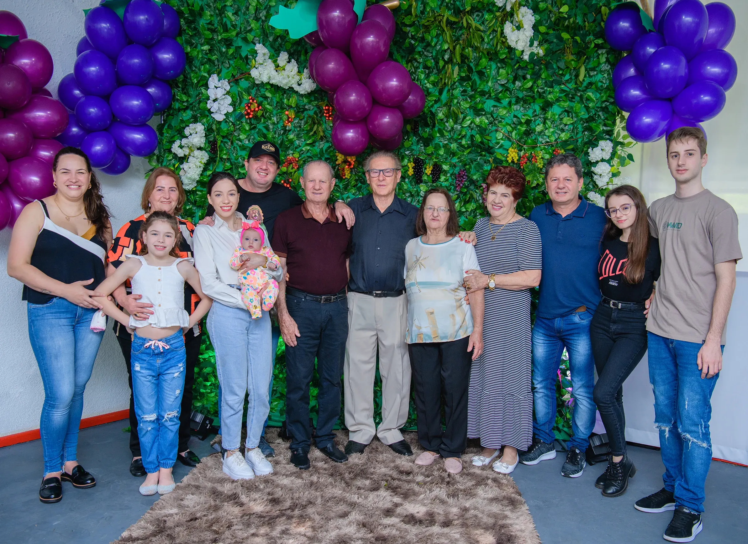 Um grupo familiar de catorze pessoas, de bebês a idosos, posa em frente a uma parede verde decorada com folhas e uvas roxas, com a maioria das mulheres à esquerda e os homens à direita.
