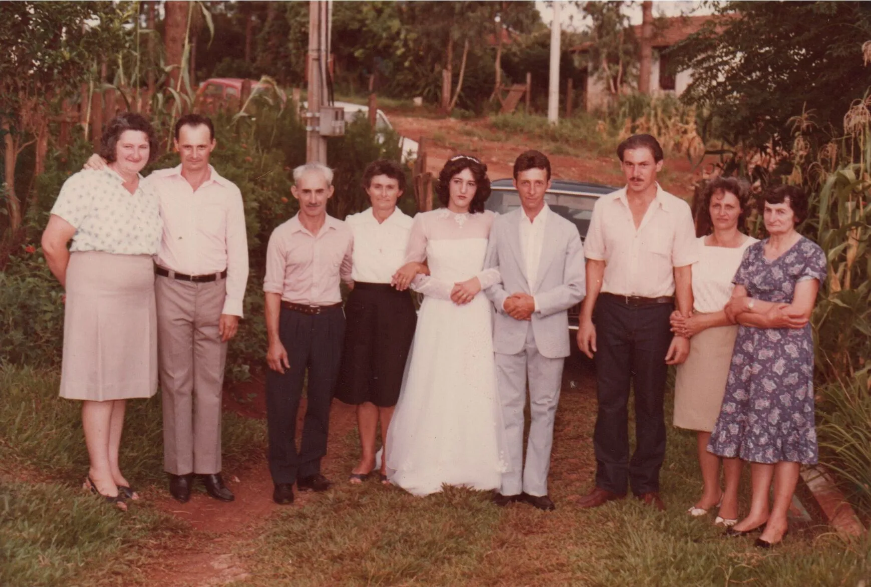 Uma noiva e um noivo estão parados no centro, sorrindo para a câmera, entre dois grupos de pessoas em um casamento ao ar livre, com árvores e grama ao fundo. (Original)