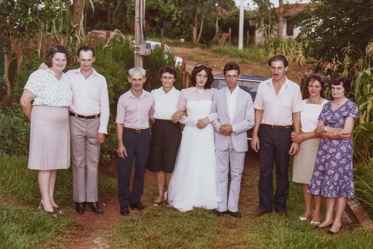 Uma noiva e um noivo estão parados no centro, sorrindo para a câmera, entre dois grupos de pessoas em um casamento ao ar livre, com árvores e grama ao fundo.