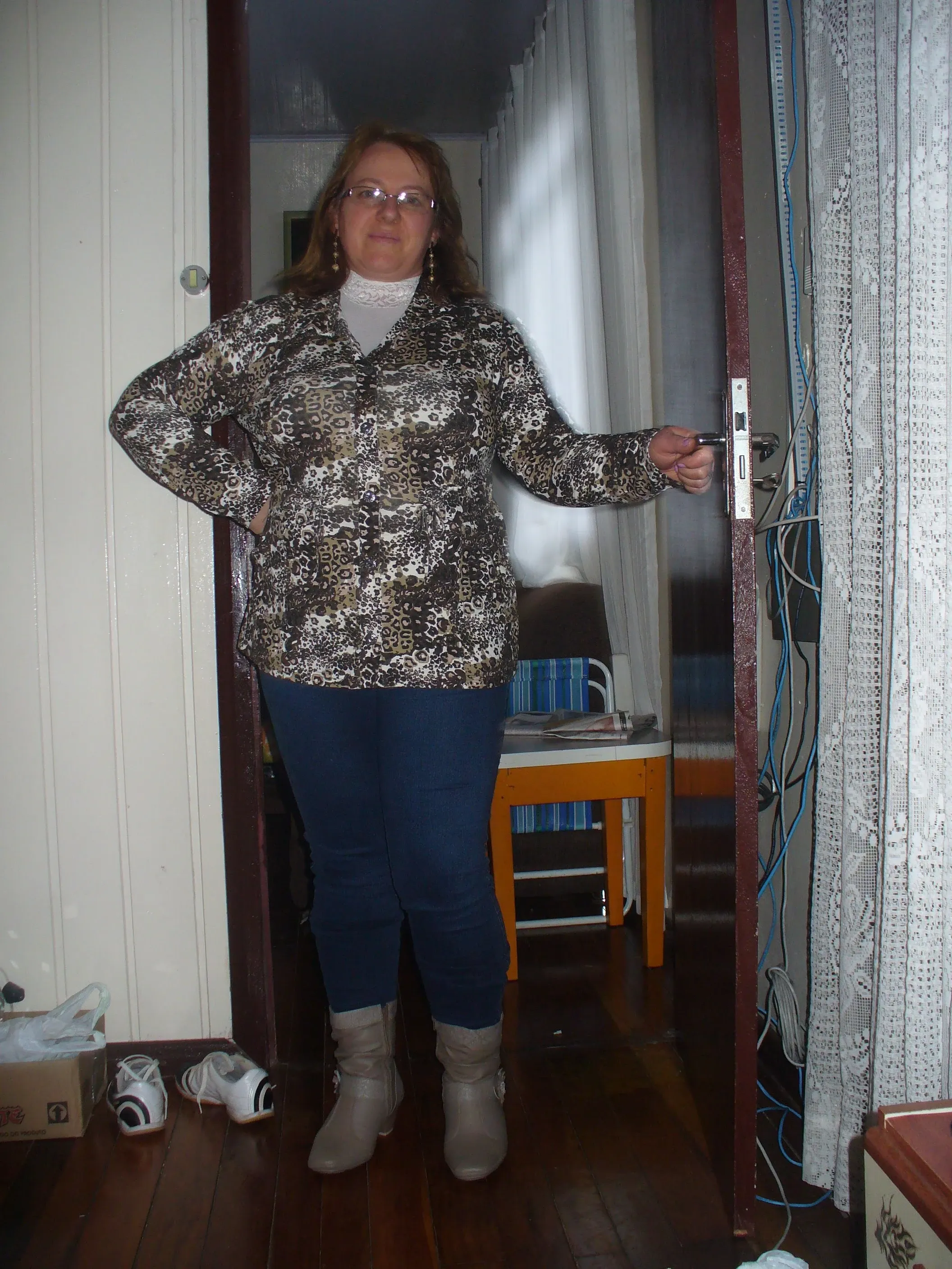 Uma mulher de meia-idade, com cabelos ruivos, óculos e um casaco estampado de oncinha sobre uma blusa de renda branca, posa com a mão no quadril e a outra segurando uma porta aberta, em um quarto com cortinas rendadas e calçados no chão.