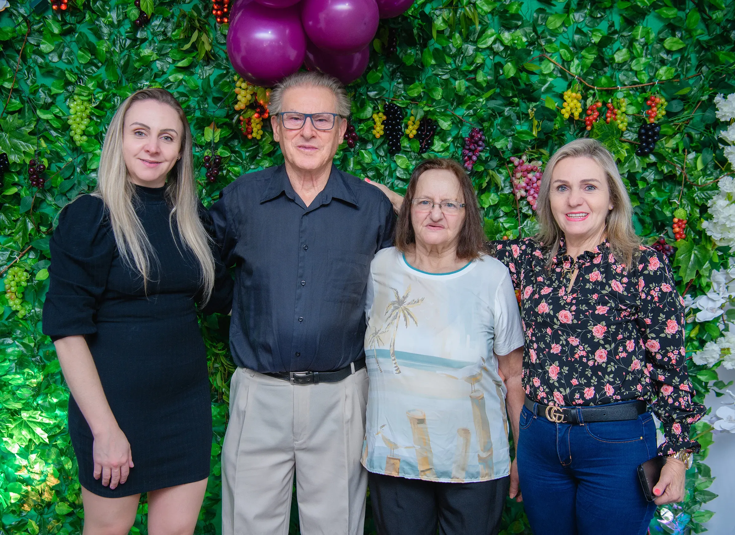 Quatro pessoas, duas mulheres mais jovens e um casal de idosos, posam de pé e sorriem para a câmera, tendo como fundo uma parede de folhas verdes adornada com cachos de uvas e flores brancas.