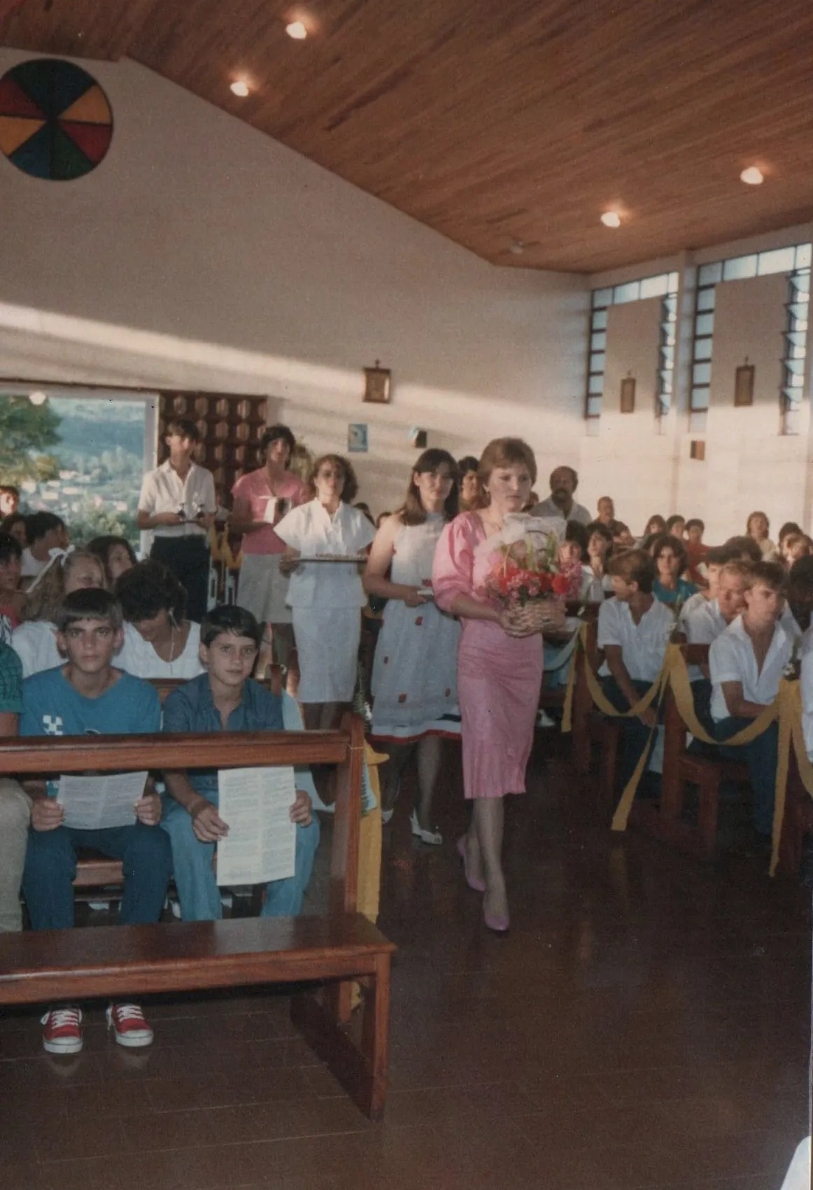 Mulher de vestido rosa com um buquê de flores caminha por uma igreja, enquanto pessoas se sentam nos bancos de madeira e assistem.