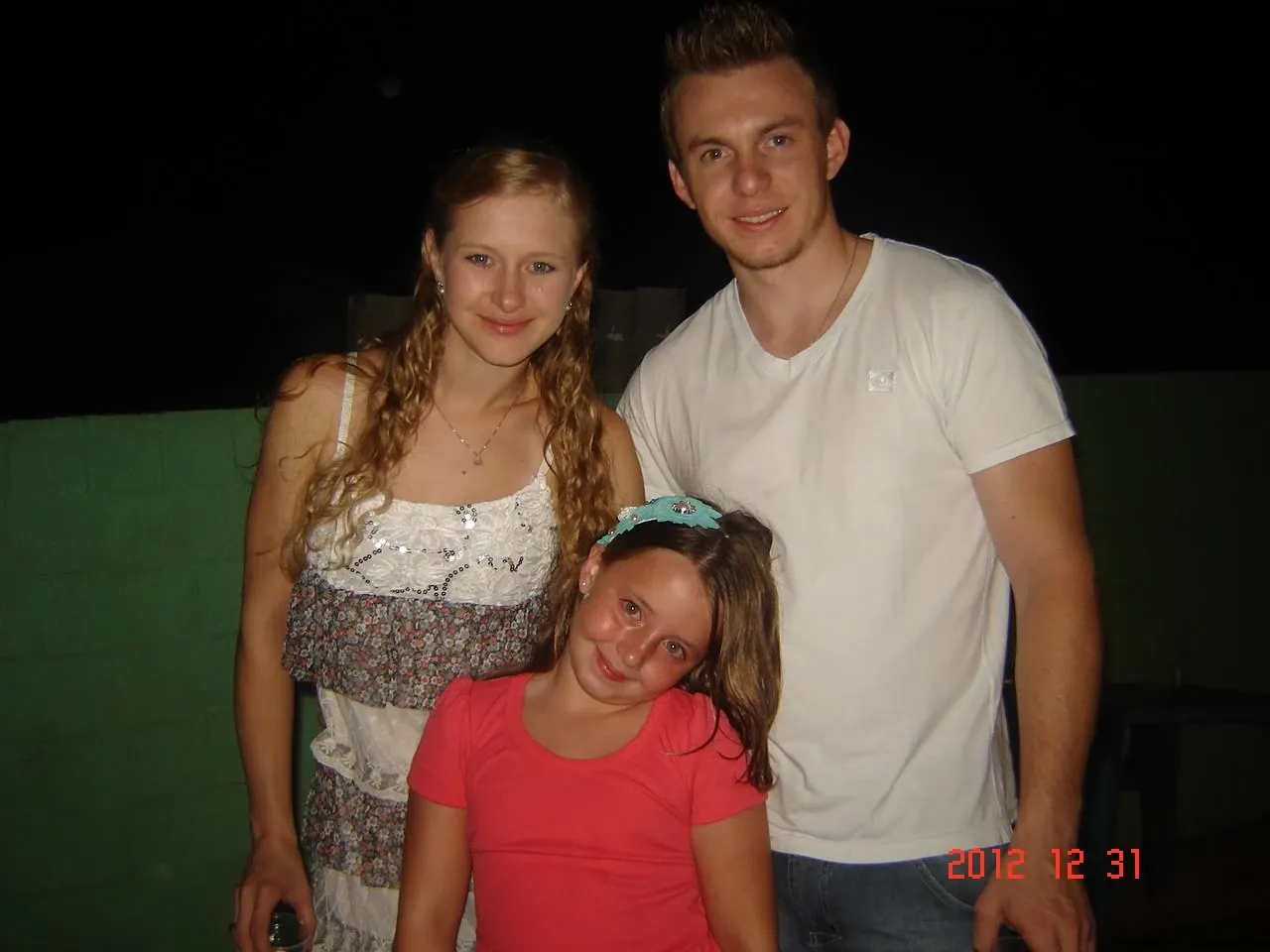 Mulher de cabelos loiros e um homem de cabelos castanhos claros sorrindo ao lado de uma menina de cabelos castanhos com uma tiara azul, todos com roupas casuais, em um ambiente escuro com iluminação fraca.