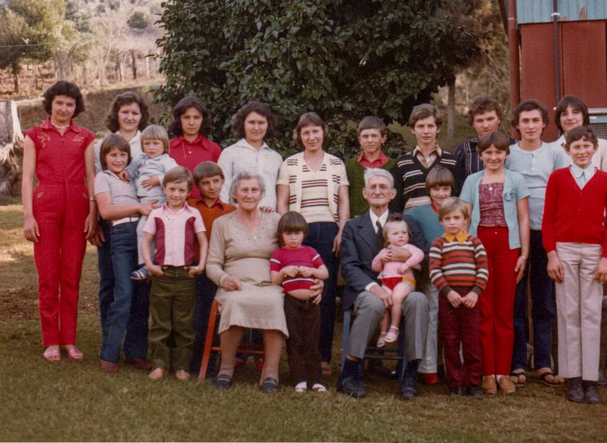 Um grupo grande e multigênero posa para uma foto de família ao ar livre, com a maioria dos membros do grupo sorrindo.