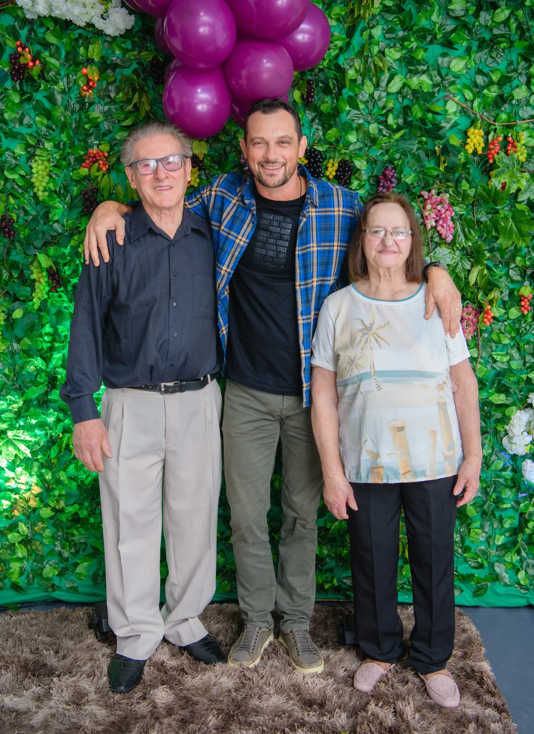 Um homem de camisa xadrez azul e camiseta preta abraça um homem idoso de camisa social preta e calças cinzas e uma mulher idosa de blusa branca estampada e calças pretas, em frente a uma parede coberta de folhagens verdes e uvas.