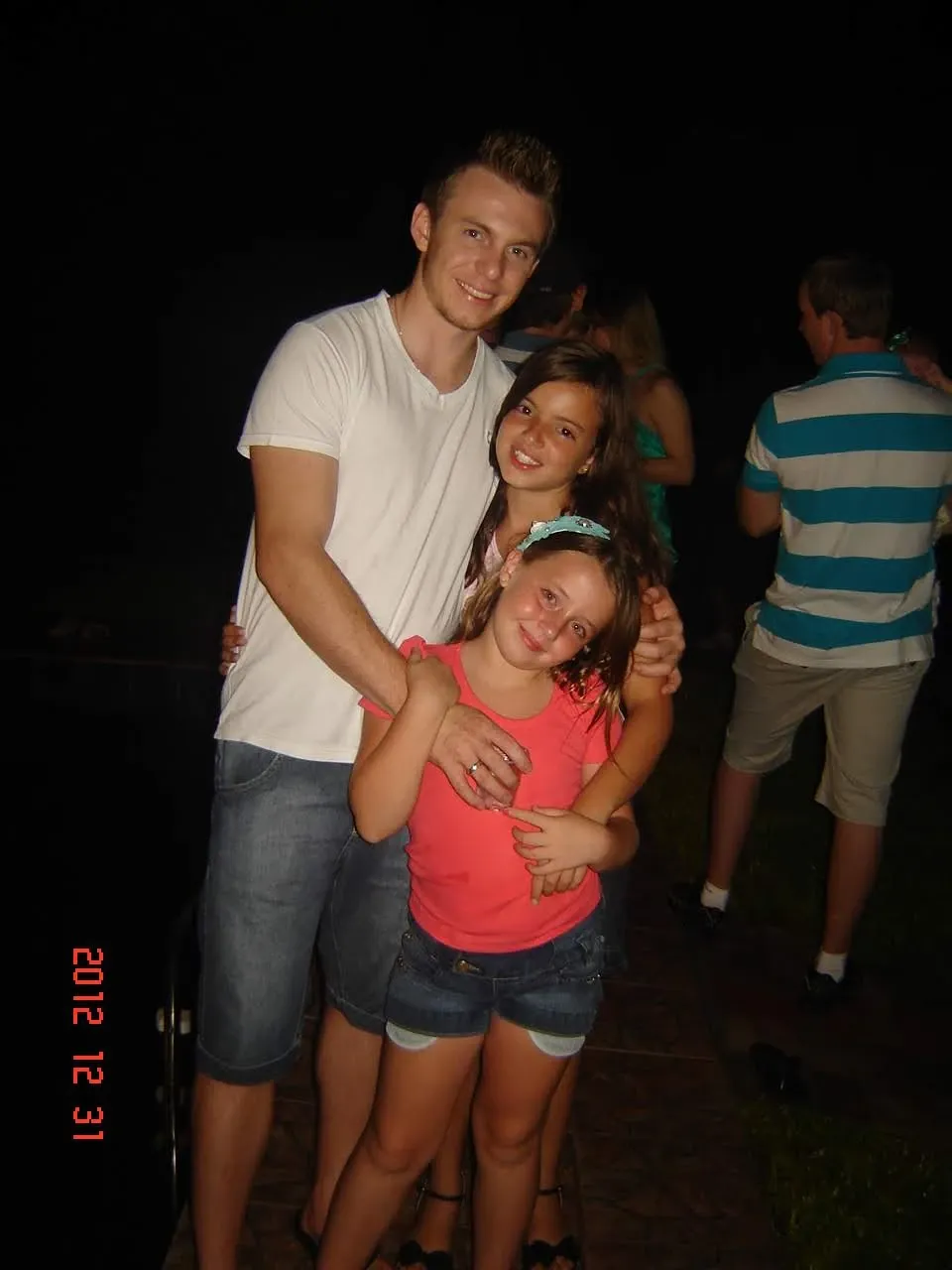 Um jovem adulto, vestindo uma camiseta branca e shorts jeans, abraça duas jovens, ambas sorrindo, em um ambiente externo com pouca luz e outras pessoas ao fundo.