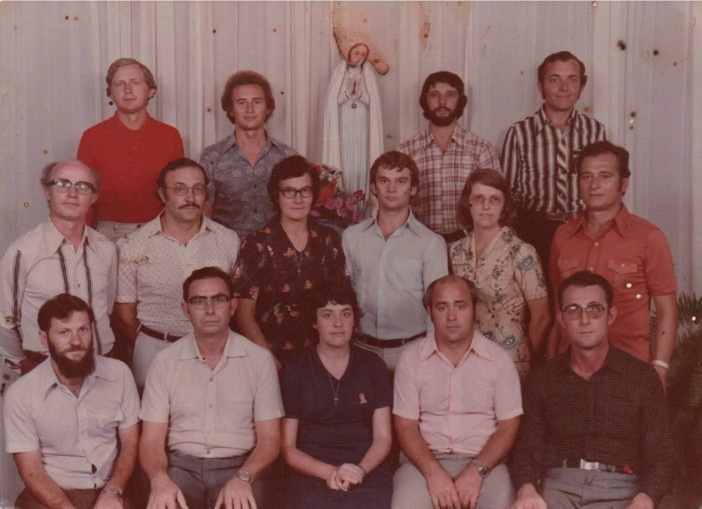 Um grupo de 15 adultos (12 homens, 3 mulheres) posando para uma foto de grupo com uma estátua da Virgem Maria no fundo. (Original)