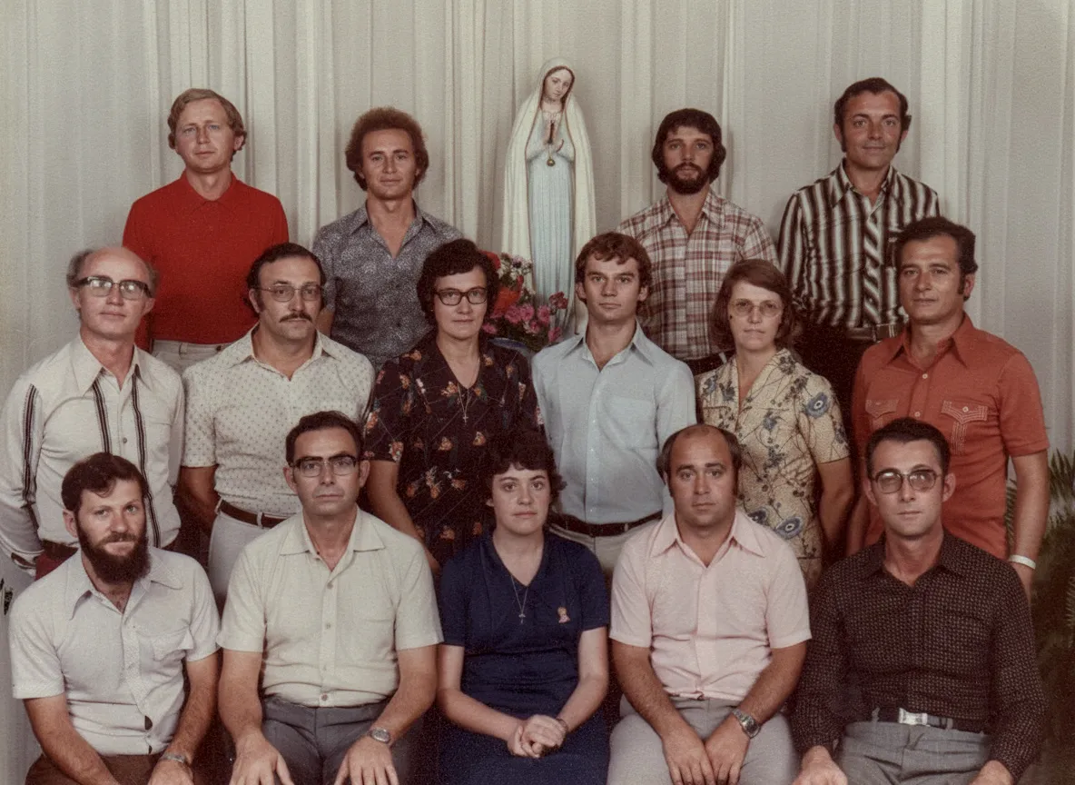 Um grupo de 15 adultos (12 homens, 3 mulheres) posando para uma foto de grupo com uma estátua da Virgem Maria no fundo.