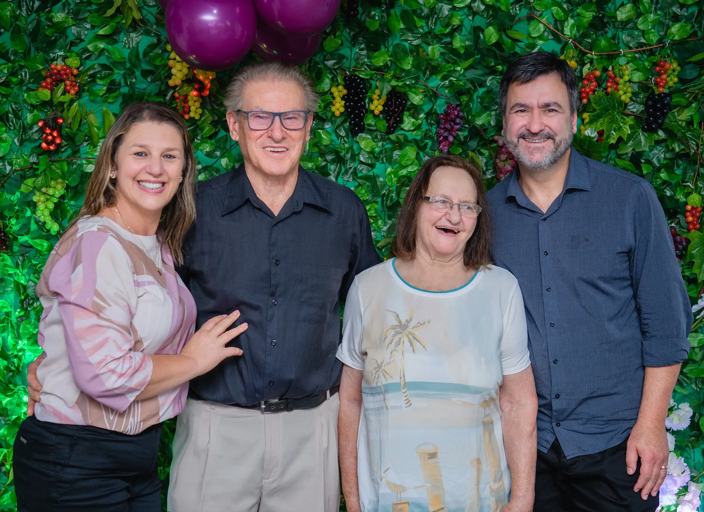 Quatro pessoas, duas mulheres e dois homens, sorriem para a câmera em frente a uma parede verde decorada com frutas e balões roxos.